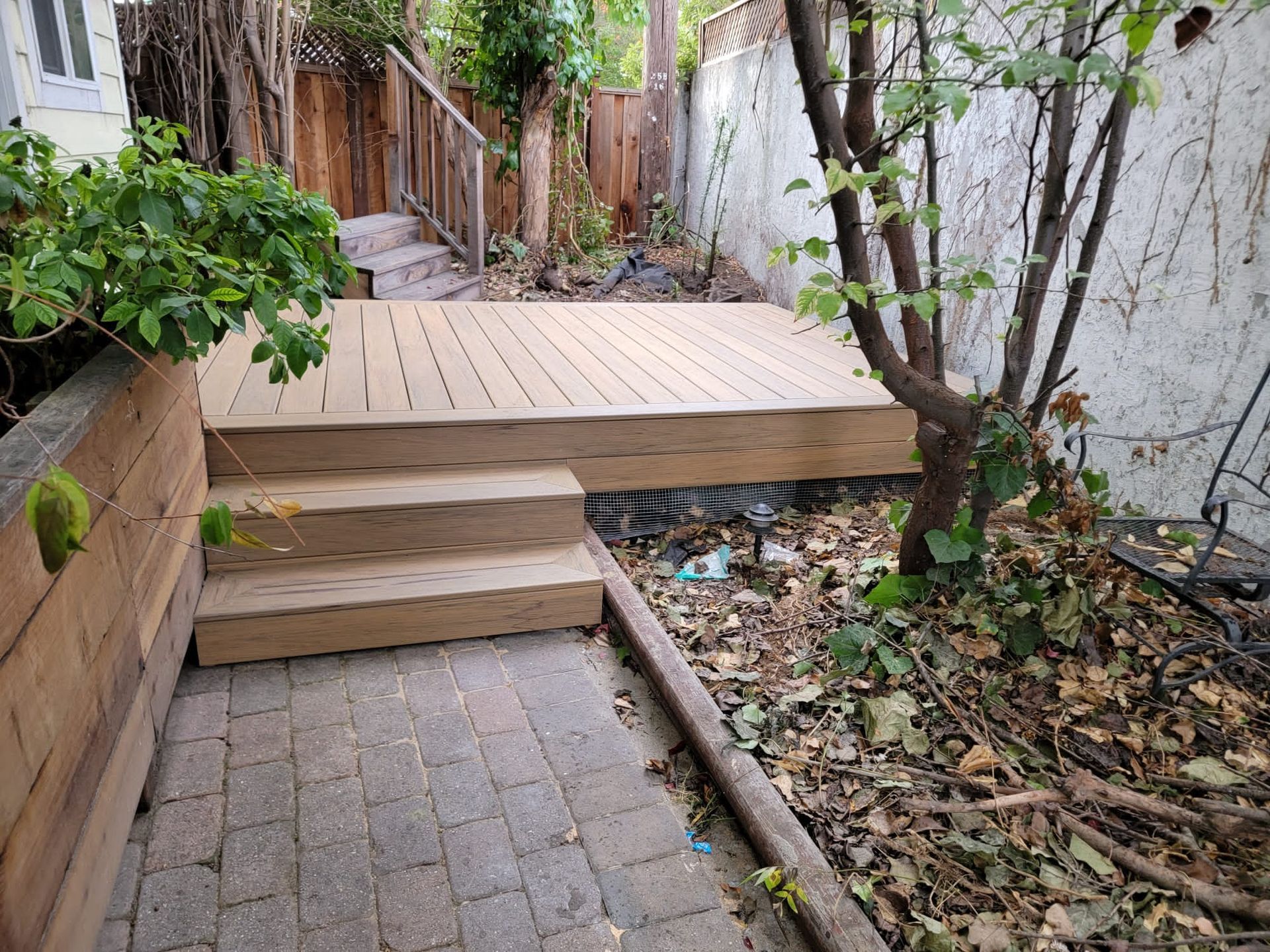 Wooden deck with steps in a backyard setting, next to a brick path and small tree.