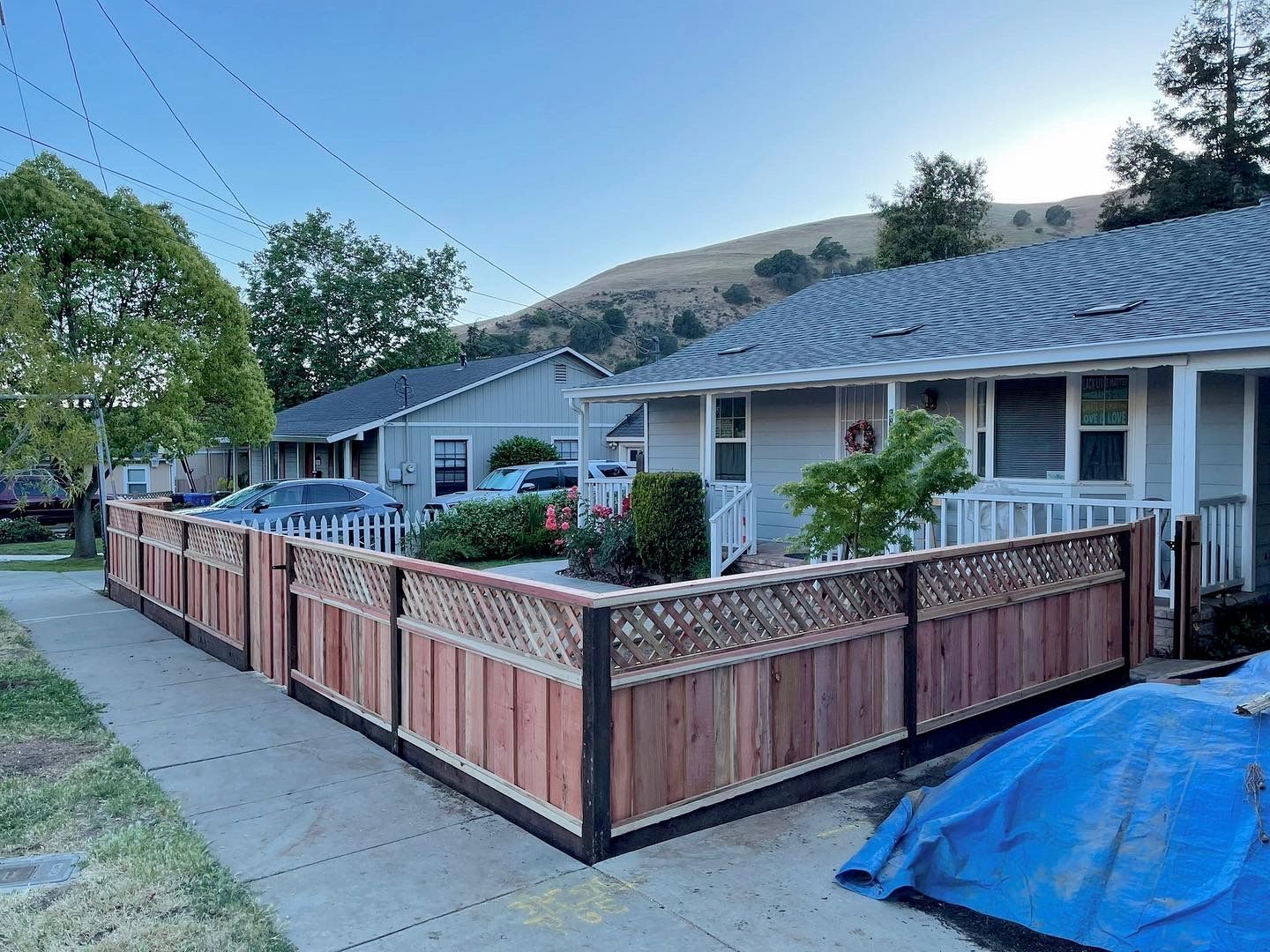 Red wood fence surrounds a light gray house. Cars are parked behind it, mountain in the background.