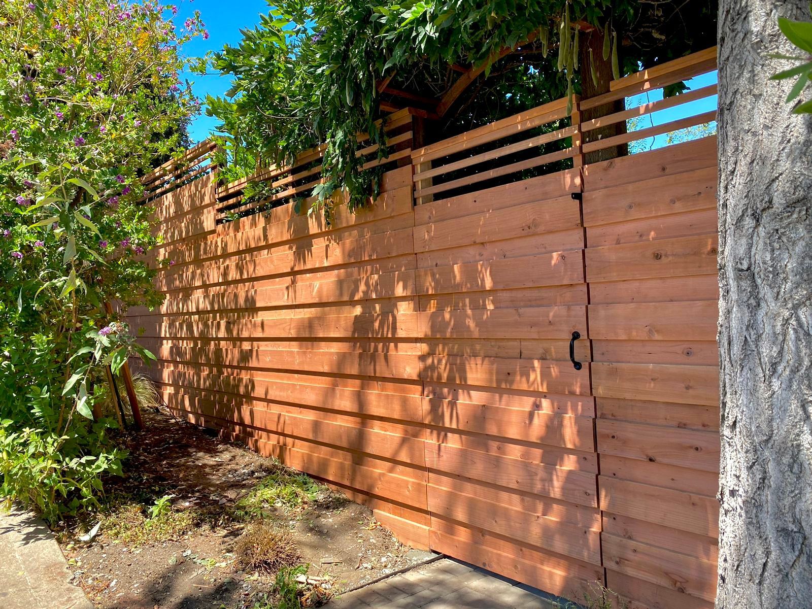 Wooden fence and gate with horizontal slats, cedar color. Green foliage surrounds.