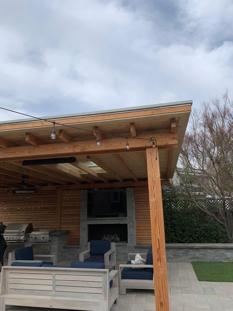 Outdoor wooden patio with a grill, fireplace, seating, and lighting under a cloudy sky.