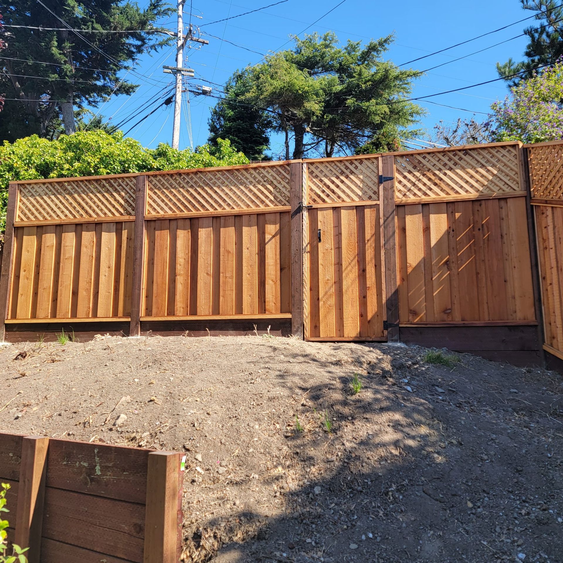 Wooden fence along a sidewalk, with a tree providing shade, under a blue sky.