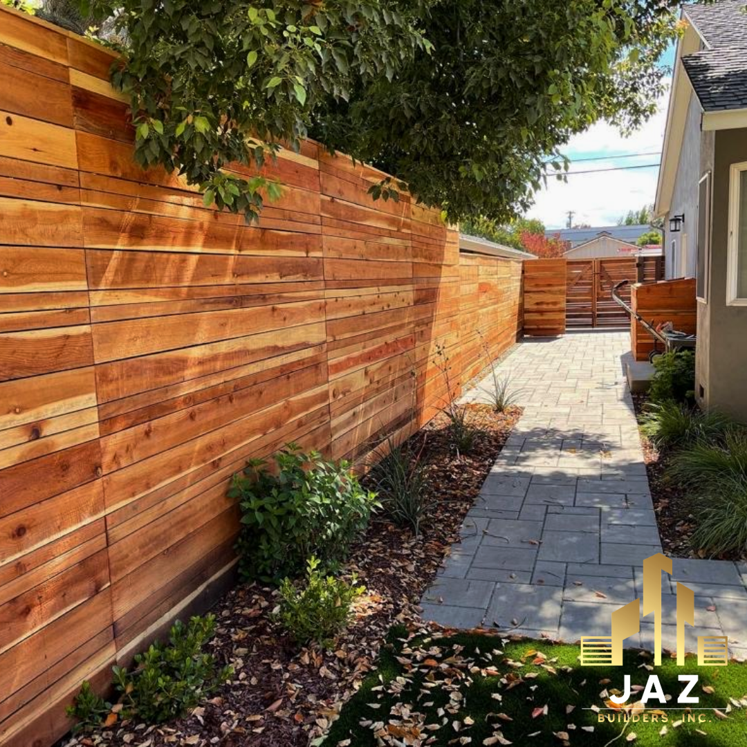 Wooden fence along a stone pathway next to a building. Sunlight casts shadows on the fence.