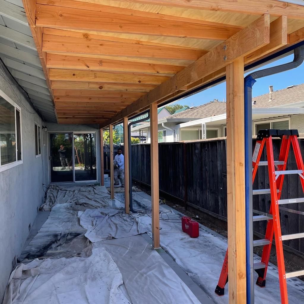 Construction of a wooden patio cover, with lumber, a person working, and a ladder.