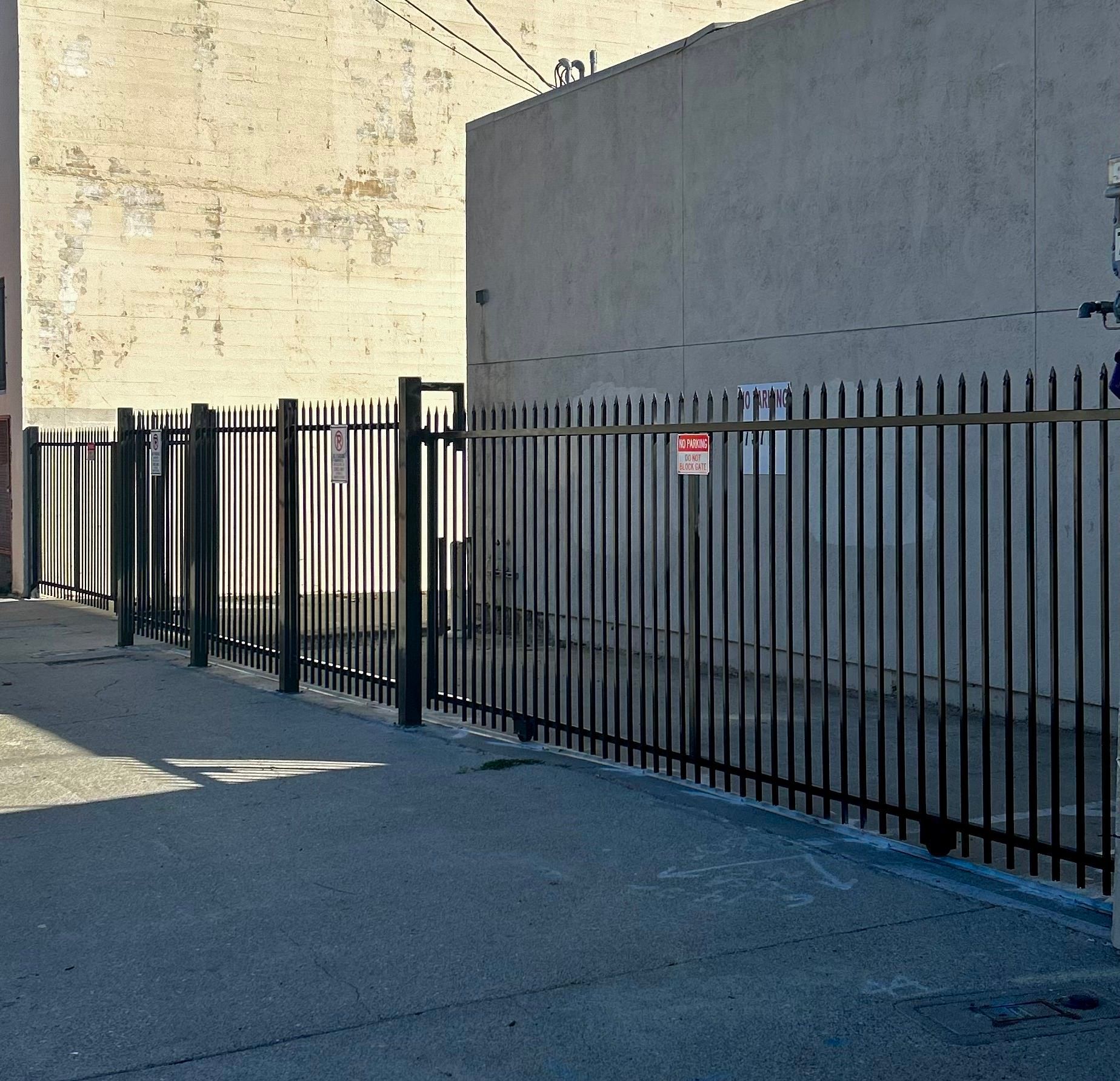 A black iron fence lines a paved lot in front of a pale, textured building wall under bright sunlight.