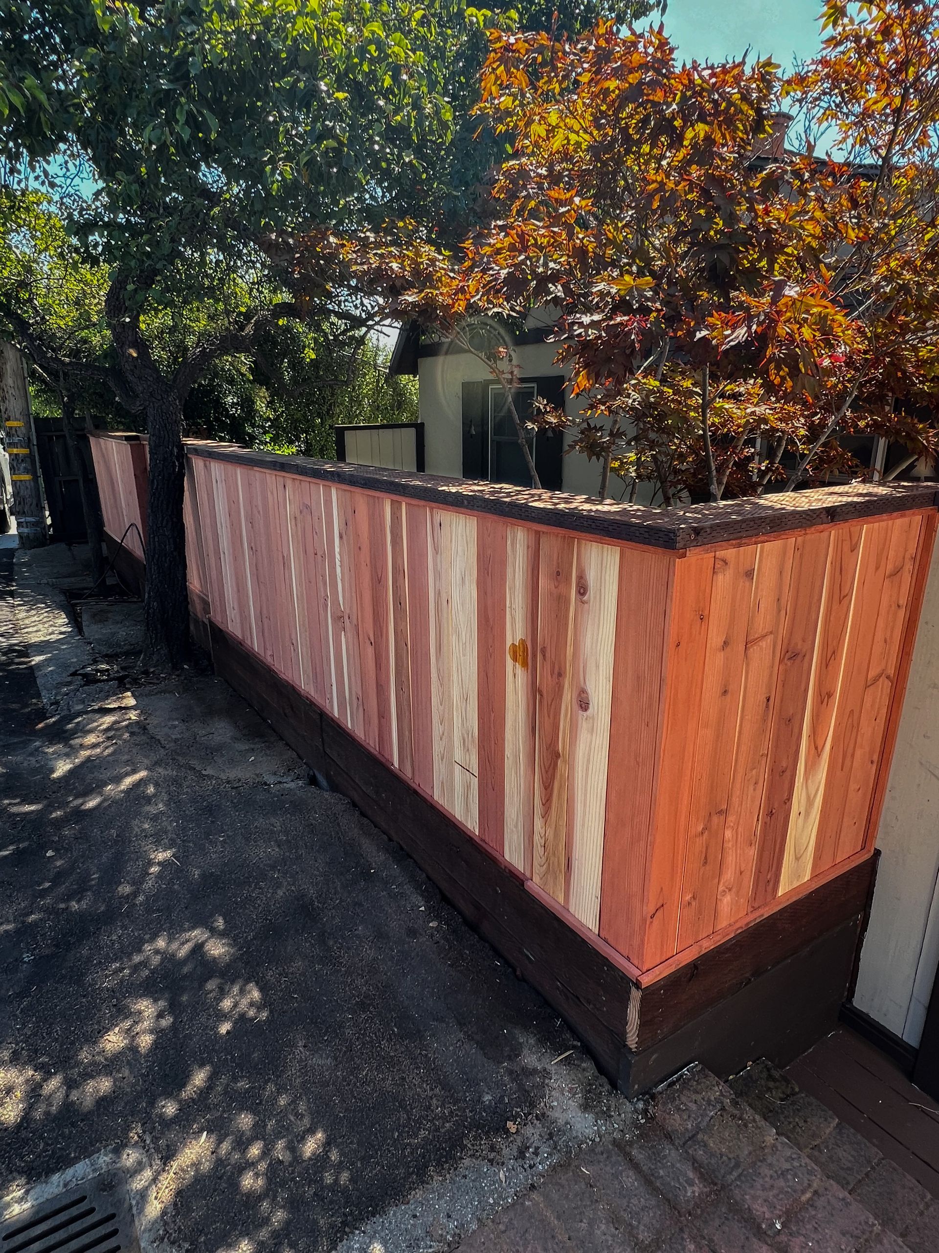 Wooden fence with varying shades of red and brown. Partially obscured by a tree, along a sidewalk.