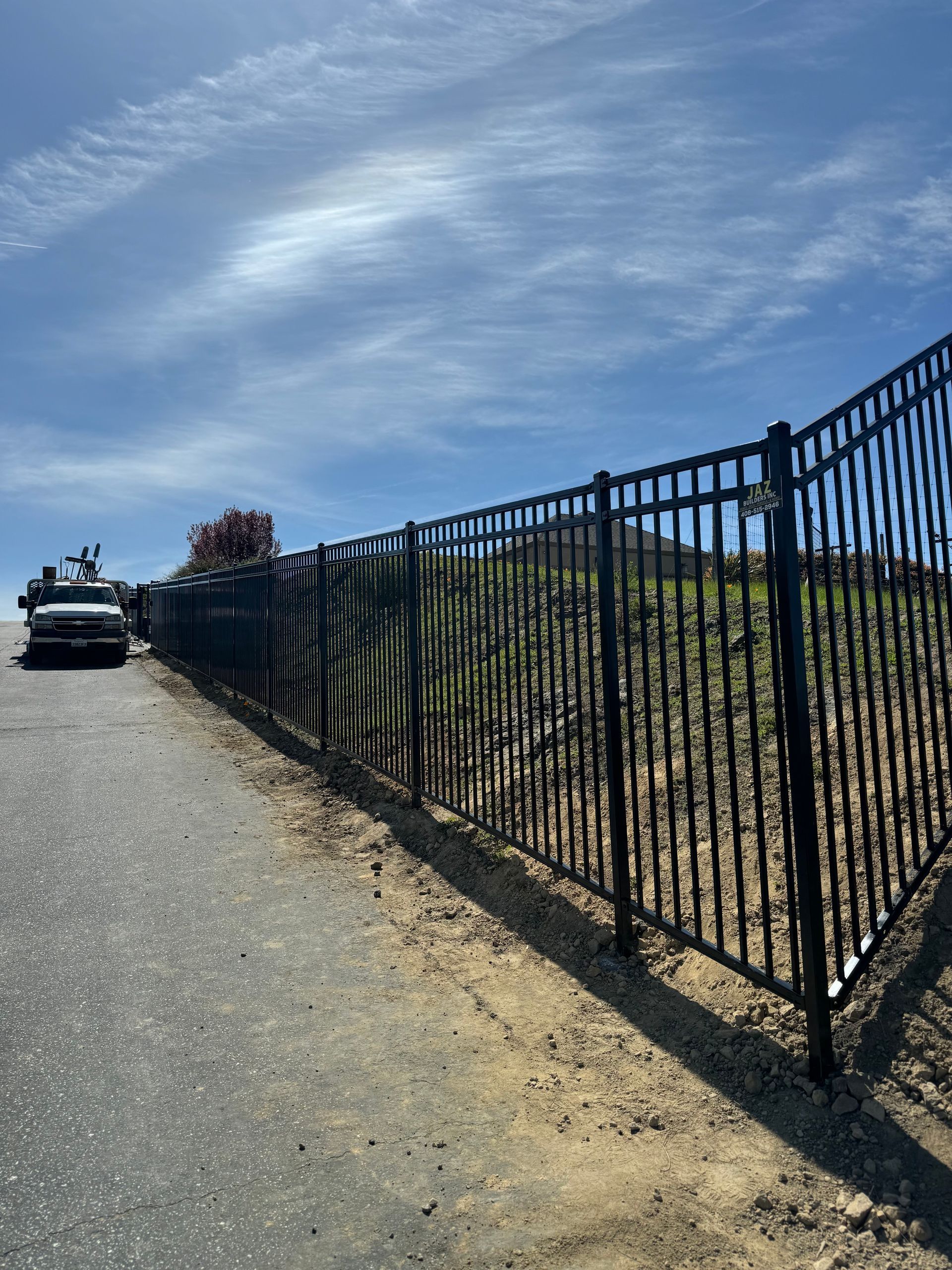 A black metal fence runs alongside a paved road under a bright blue sky with light clouds.