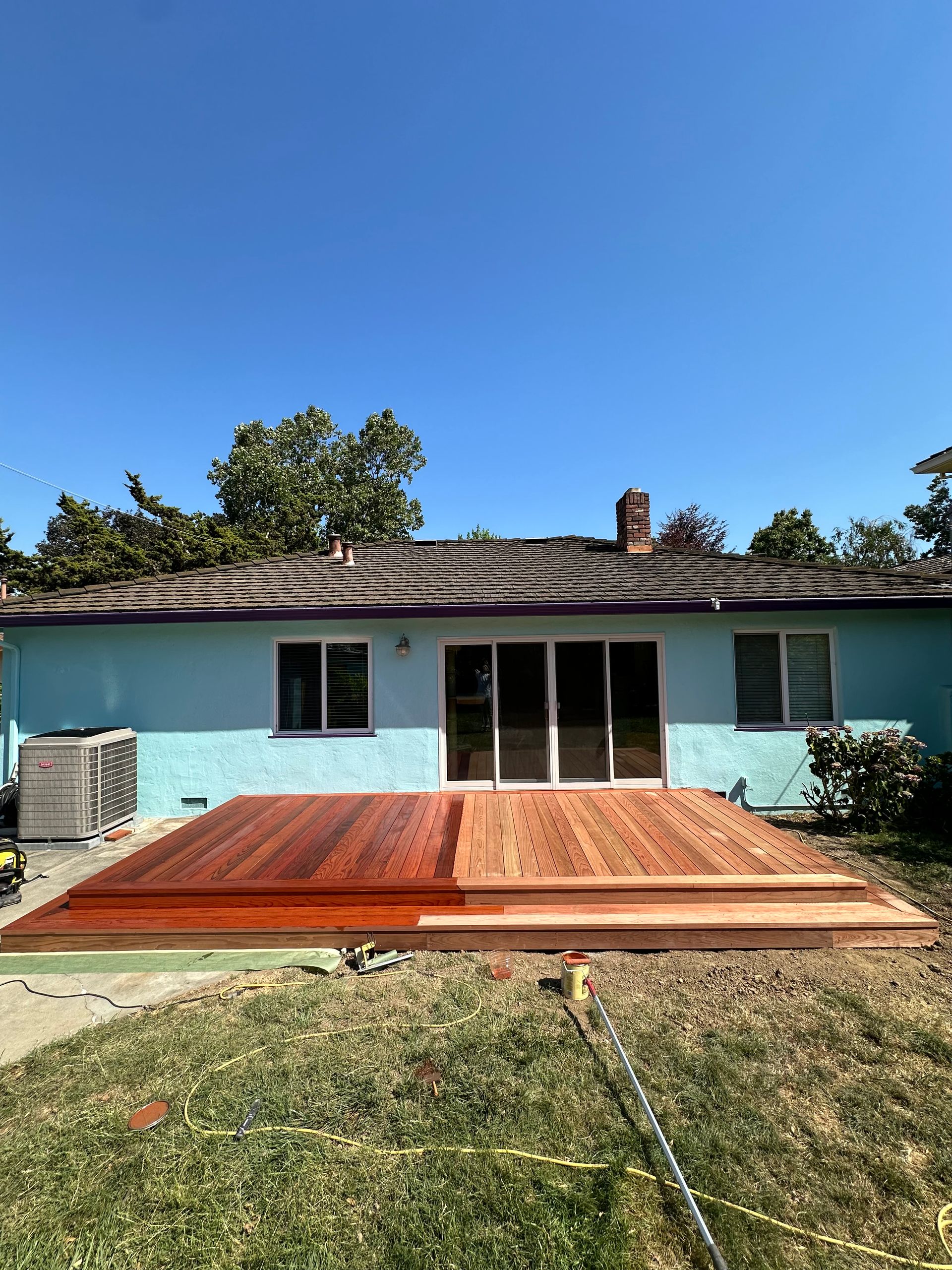 A partially stained wooden deck attached to a light blue house under a clear blue sky.