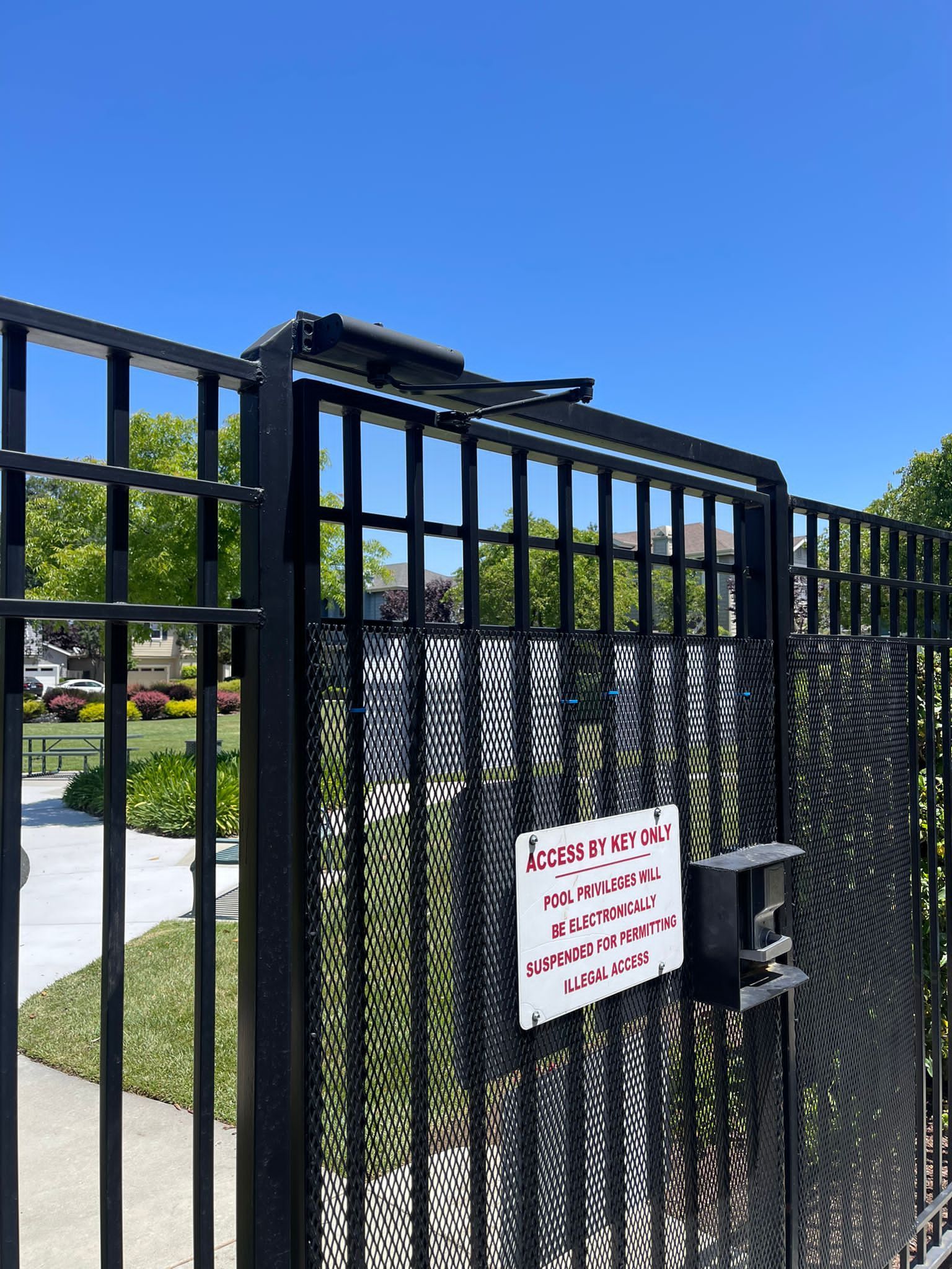 Black metal gate with a sign. Sunlight, a blue sky, and green grass are visible.