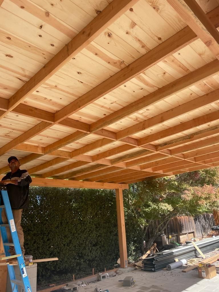 Man on ladder builds a wooden patio roof. Light wood beams and boards. Green hedge background.