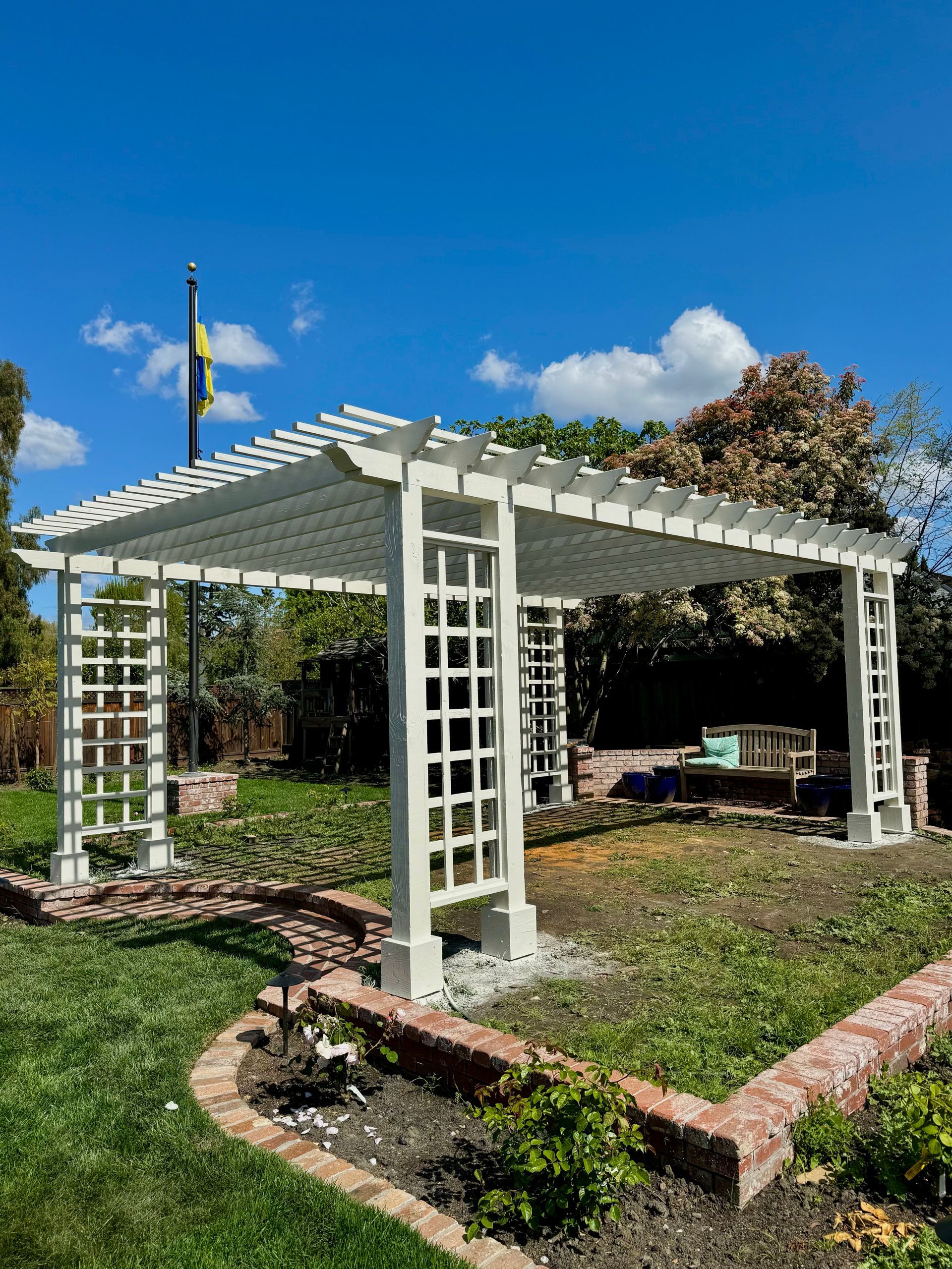 White lattice pergola in a grassy yard, surrounded by a brick border, under a blue sky.