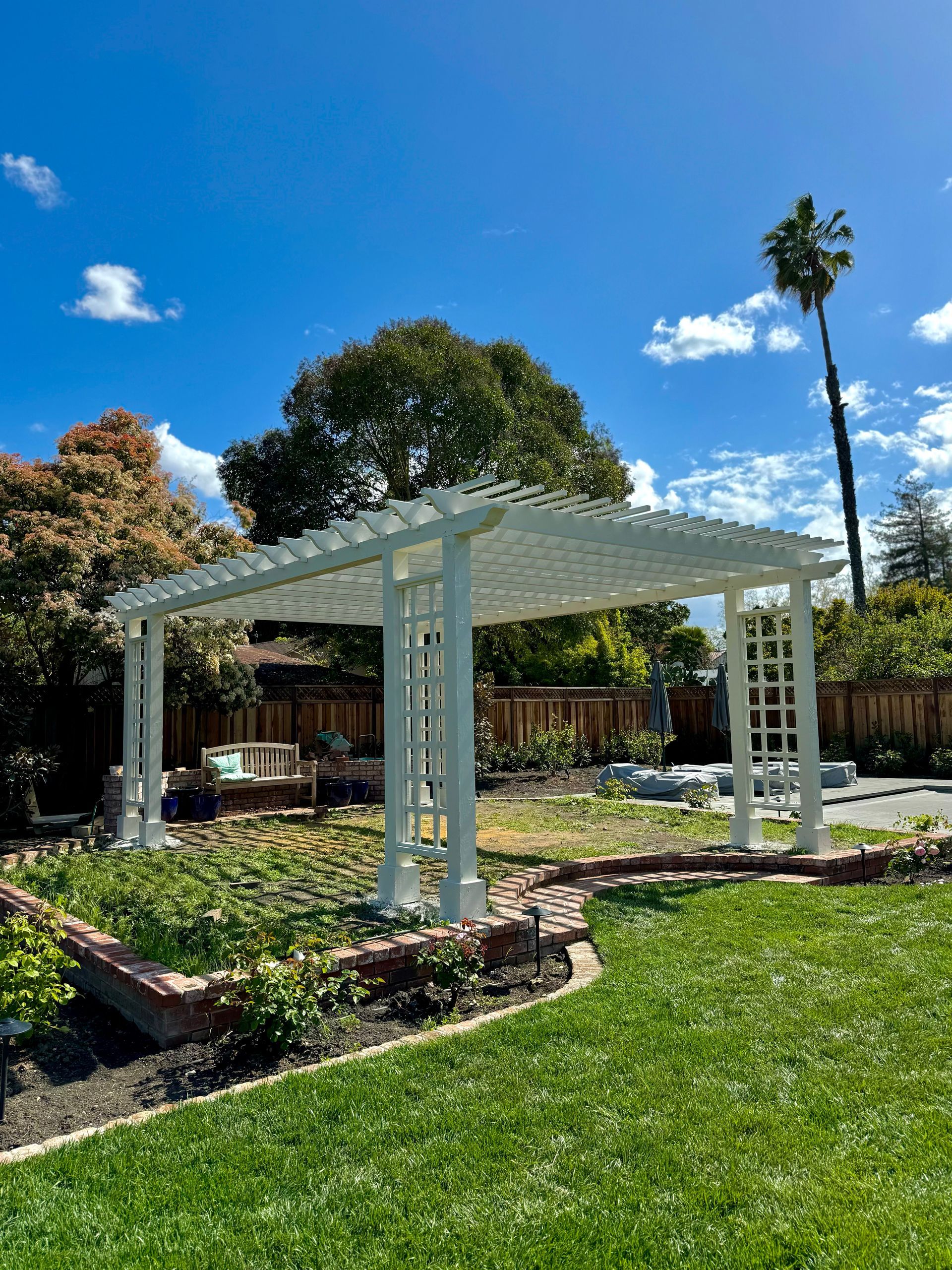 White pergola in a garden with green grass, brick border, and a blue sky.
