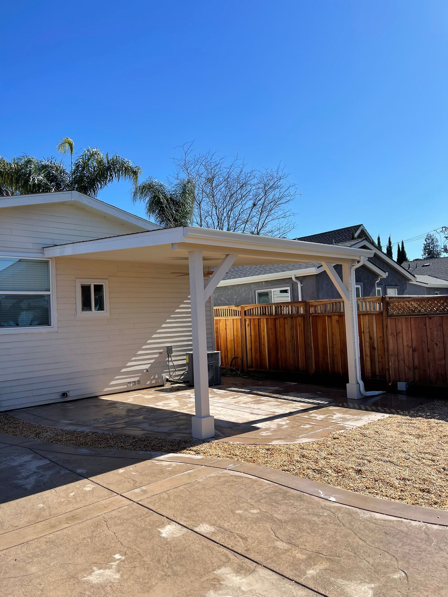 Backyard patio with wooden canopy and gravel ground, next to a house with a clear blue sky.