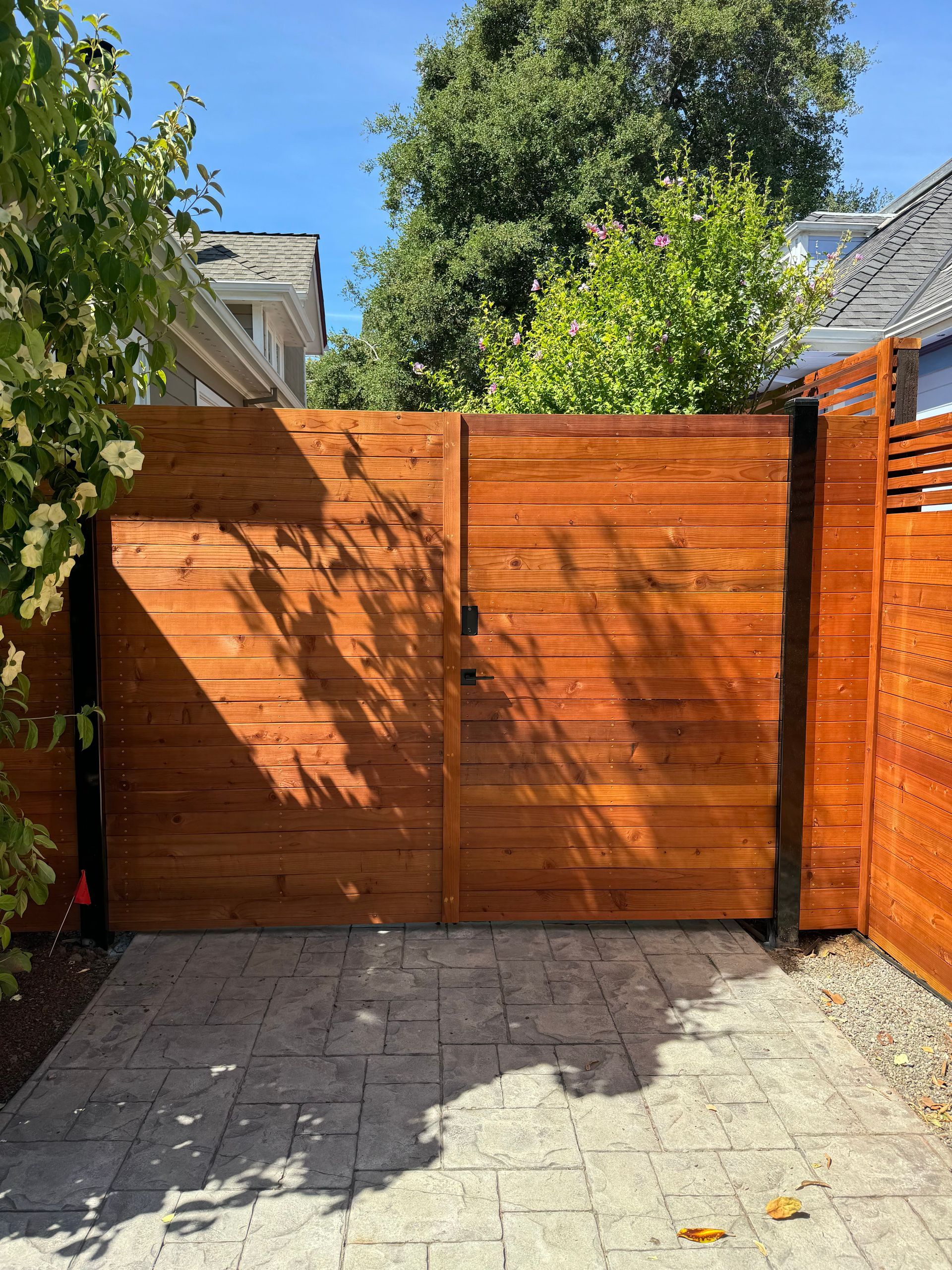 Wooden gate in a paved area, bordered by a wooden fence. Bright sunlight casts shadows.