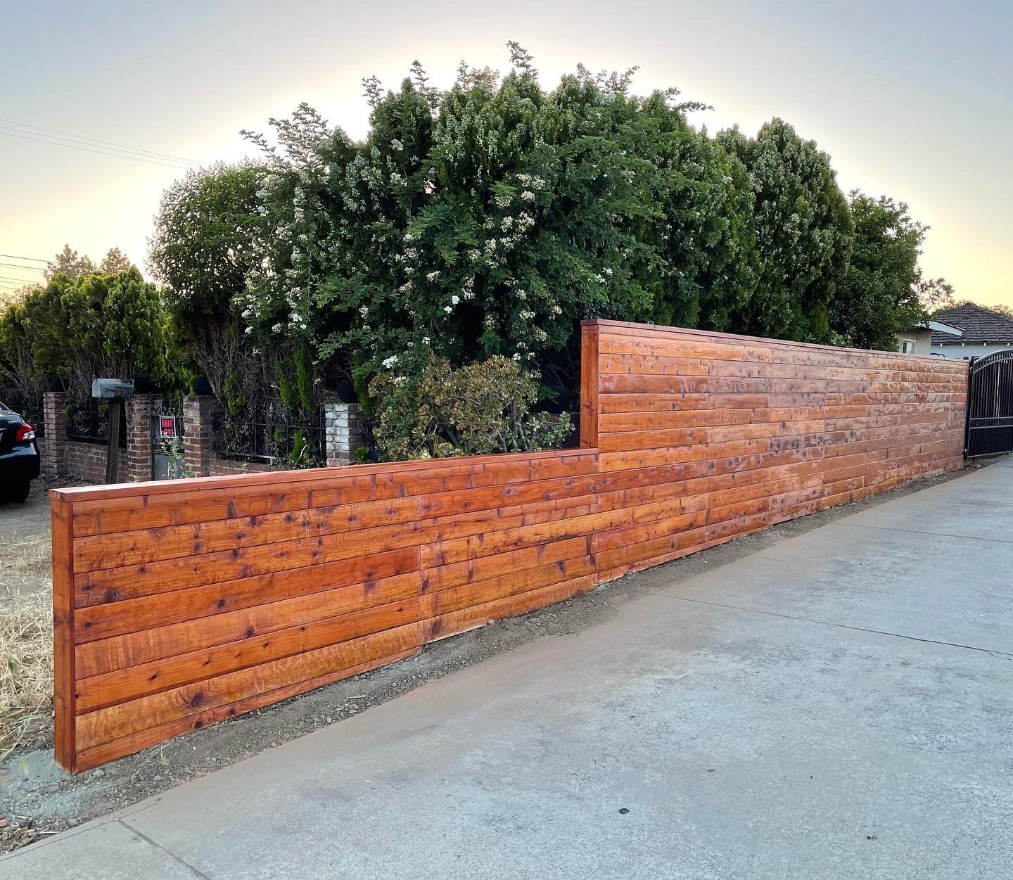 Wooden fence stained in a warm hue, bordering a driveway and lush green trees.