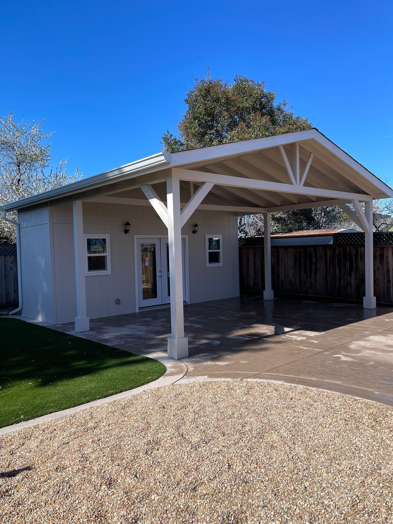 Beige building with white-trimmed windows and an attached white-beamed patio, gravel and grass in front.