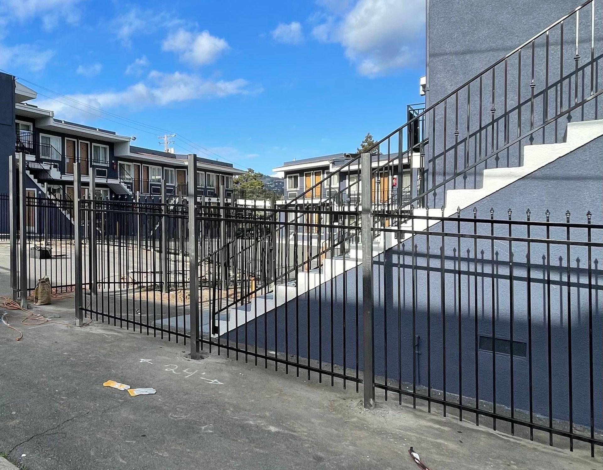 A black metal fence runs along the front of a grey residential building complex with stairs under a blue sky.