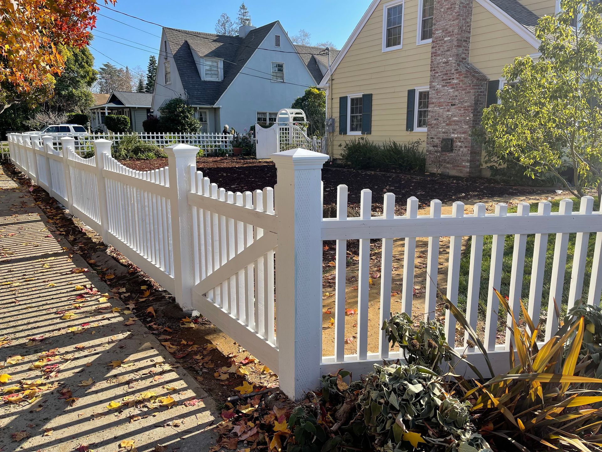 White picket fence along a sidewalk in front of houses; fall leaves scattered.