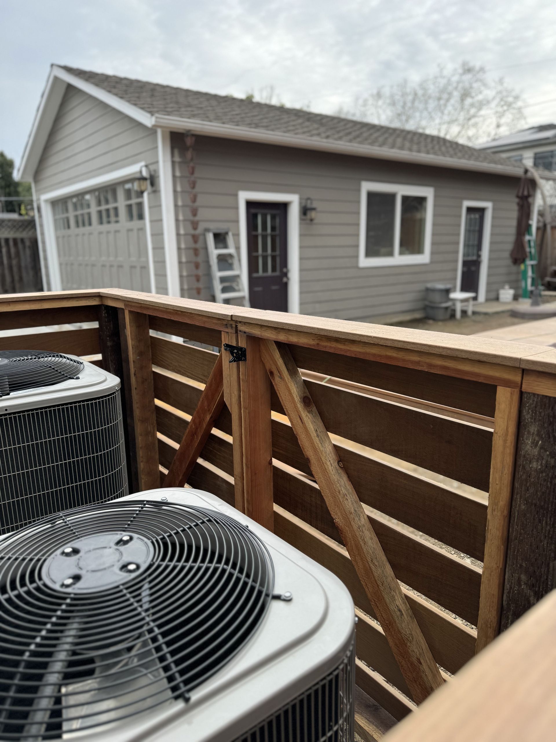 Air conditioners behind a wooden fence, small building in background with a garage door and door.