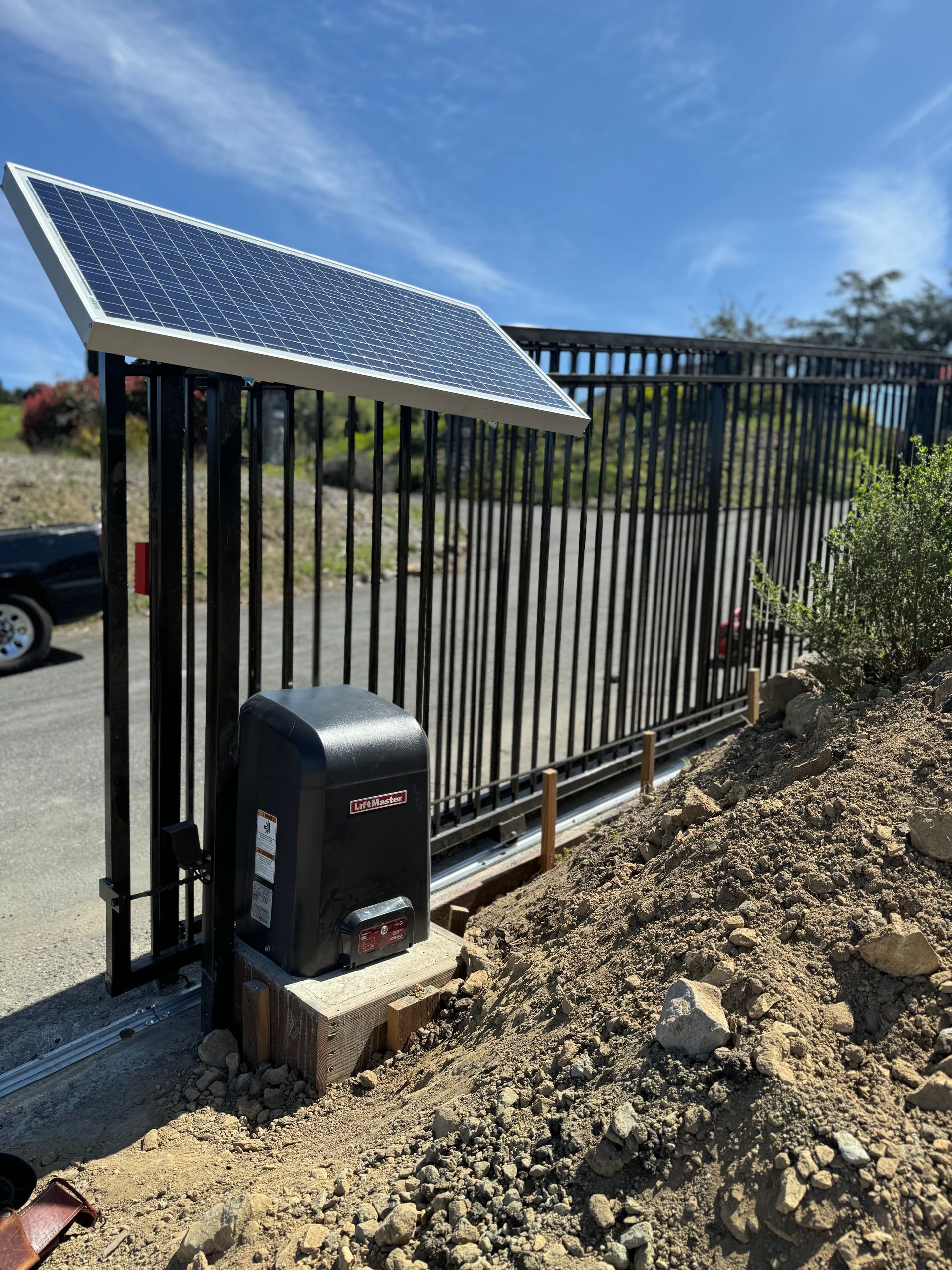 Black metal security gate with solar panel and motor on a dirt driveway.