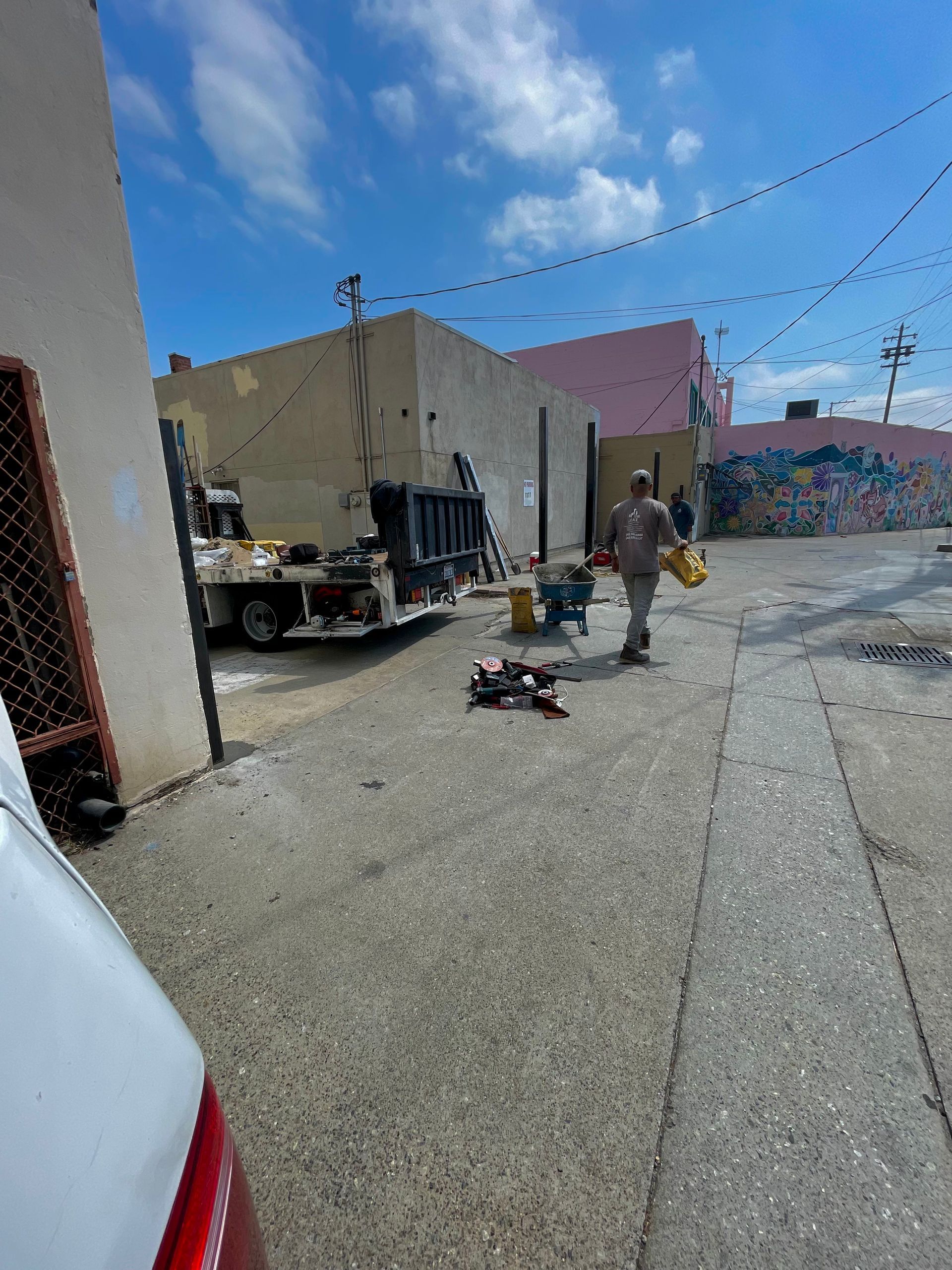 Workers near a garbage truck in an alley, one carrying items. Buildings line the street under a blue sky.