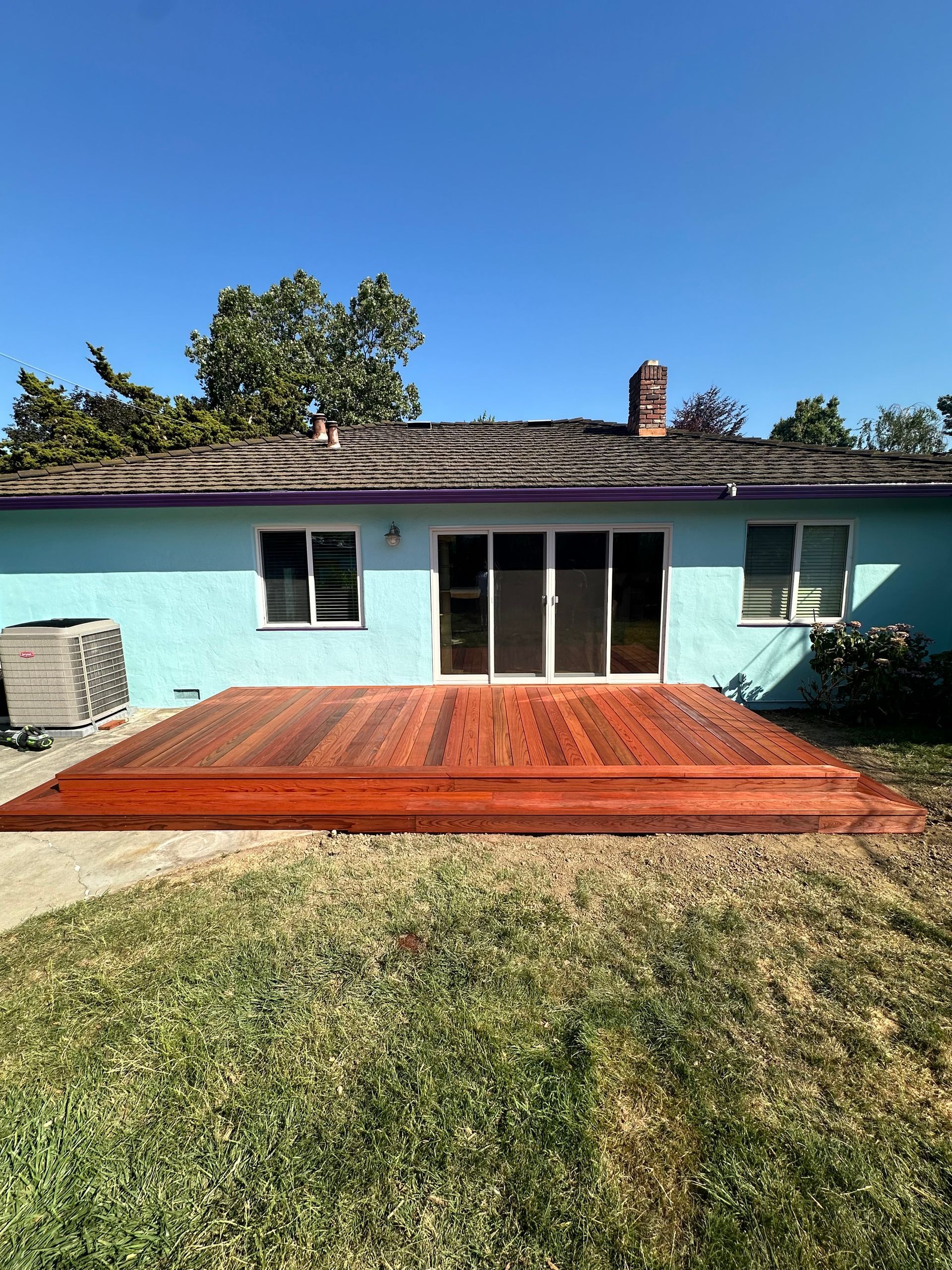 Backyard with a wooden deck, a light blue house, green grass, and a clear sky.