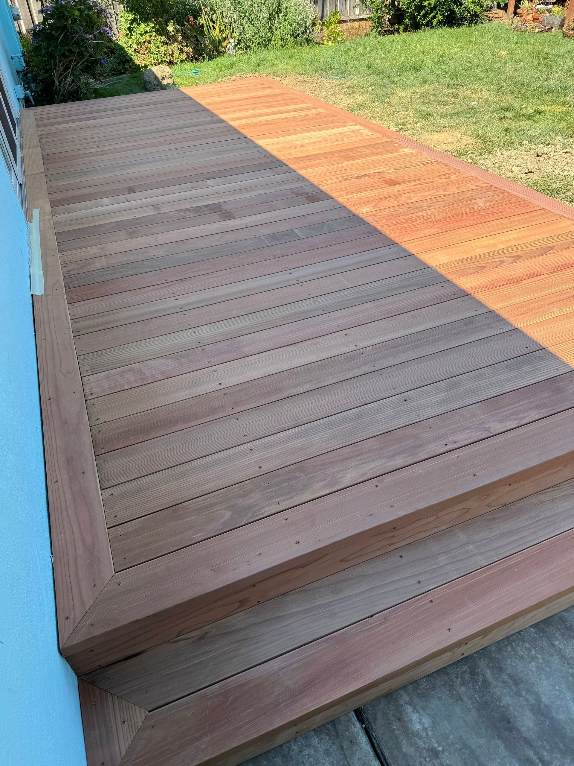 Wooden deck with two steps next to a light blue wall, on a grassy lawn.