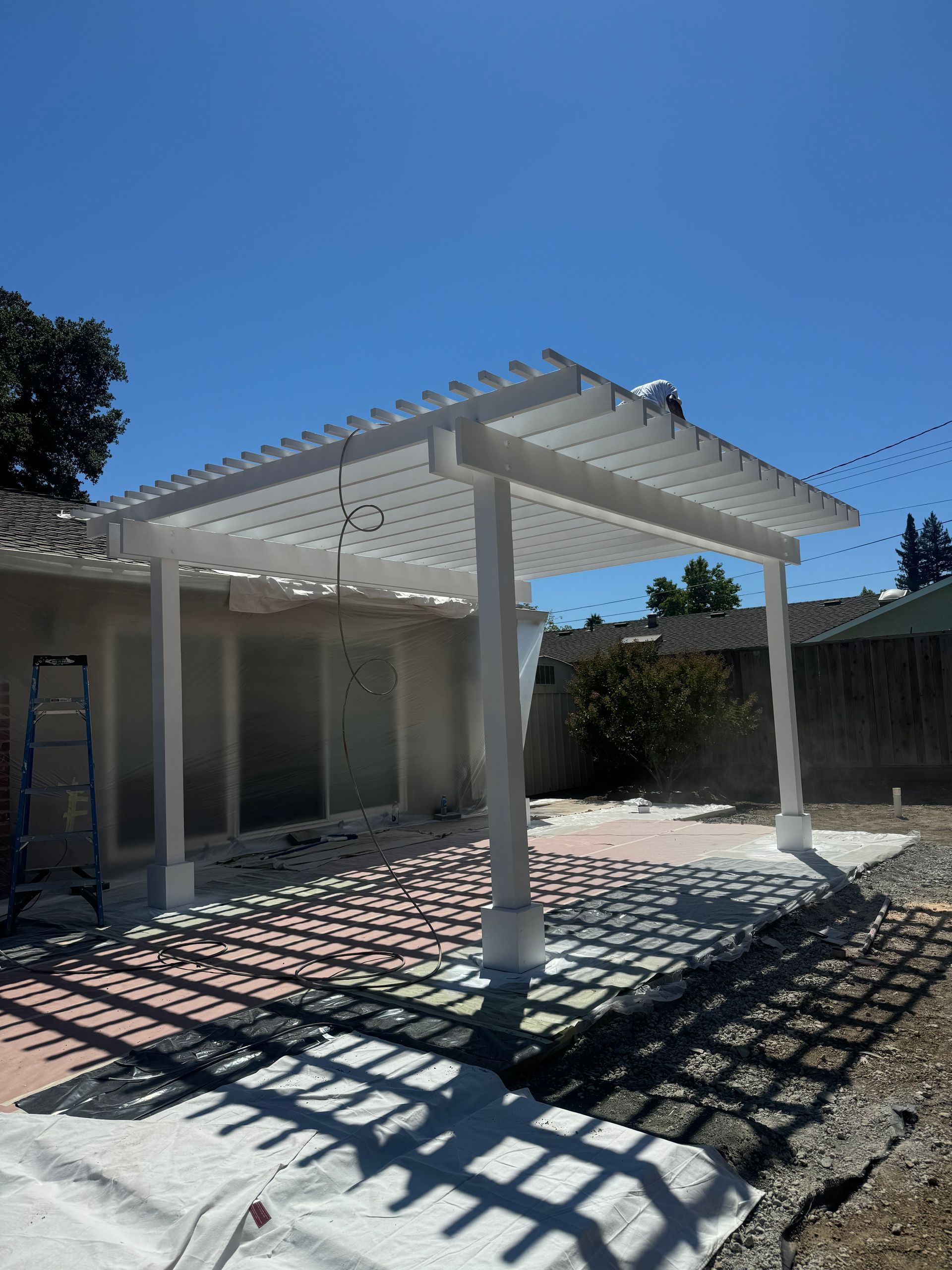 White pergola over a patio, casting shadows on the red brick and gravel ground. Bright blue sky.