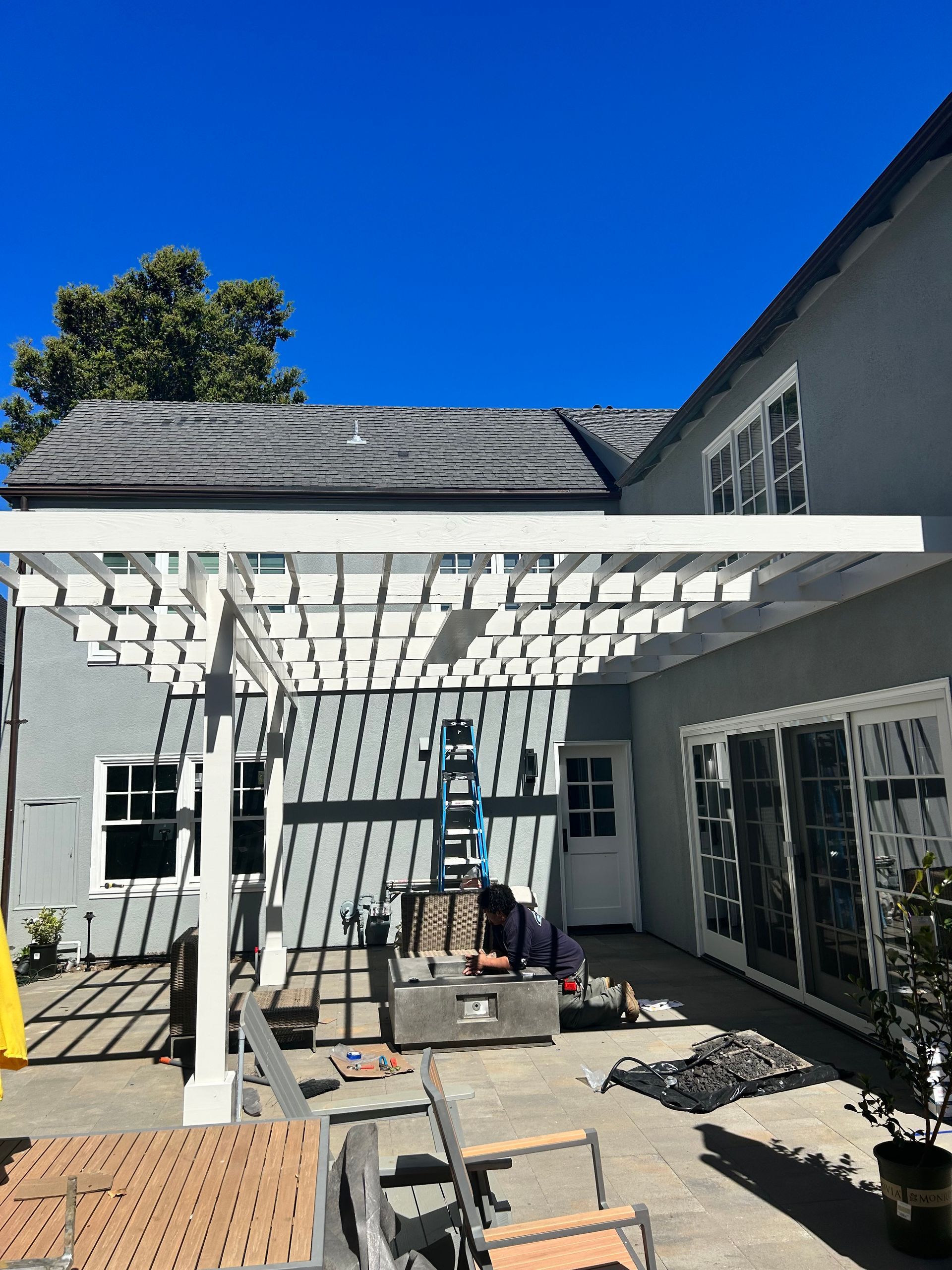 White pergola attached to a gray house, construction in progress. Blue sky overhead.