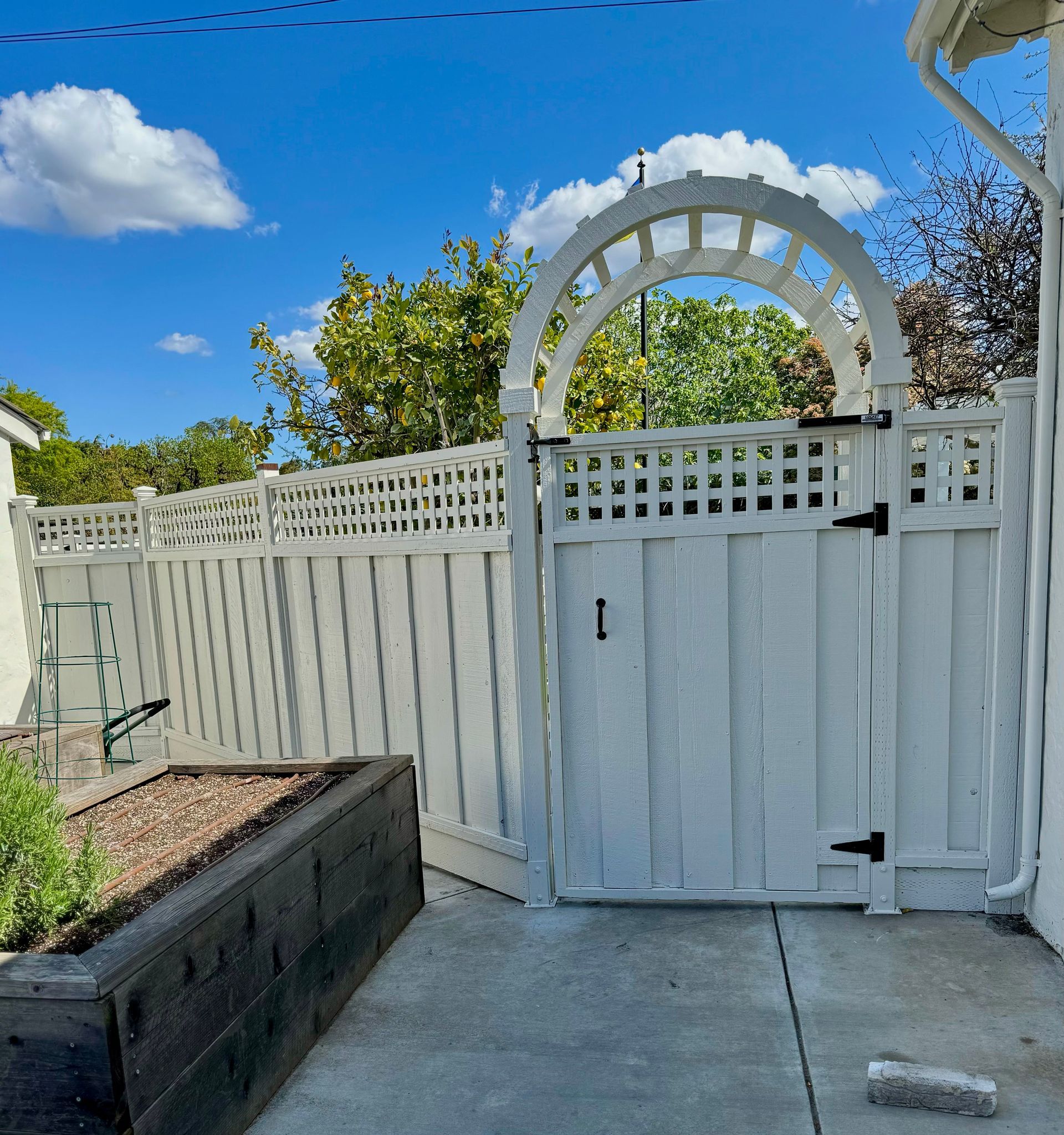 White picket fence with arched gate under a blue sky. A raised garden bed is in the foreground.