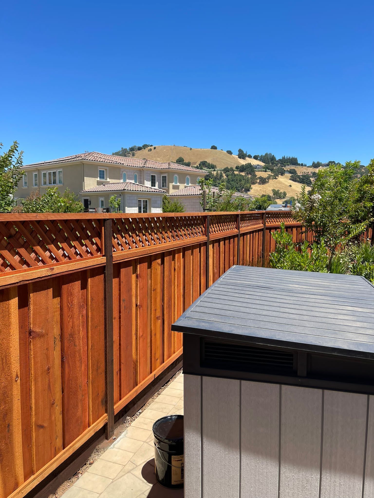 Wooden fence with decorative top, small shed, and hillside buildings under a clear blue sky.