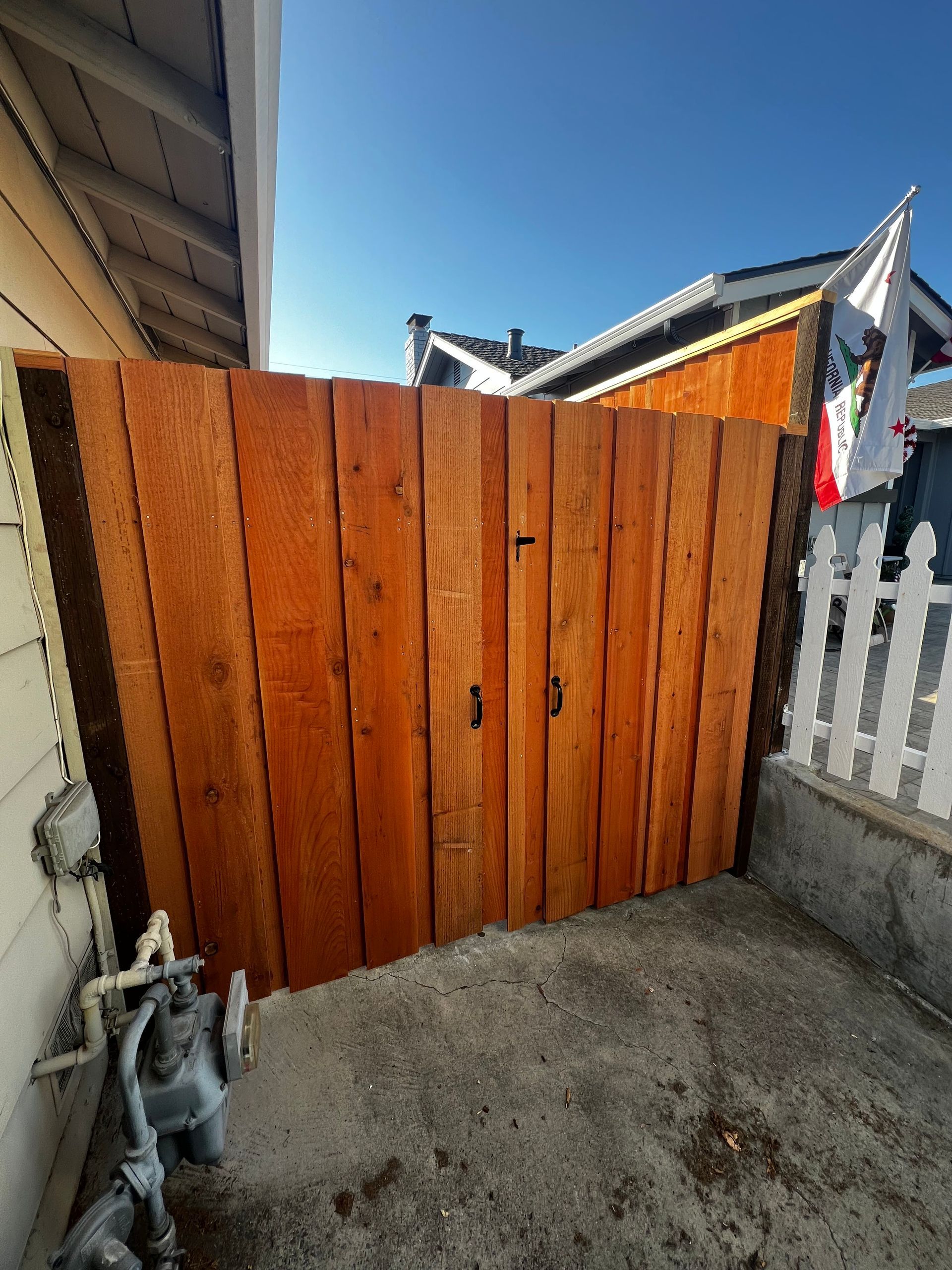Wooden gate between a building and a white picket fence, stained a reddish-brown color, set against a blue sky.