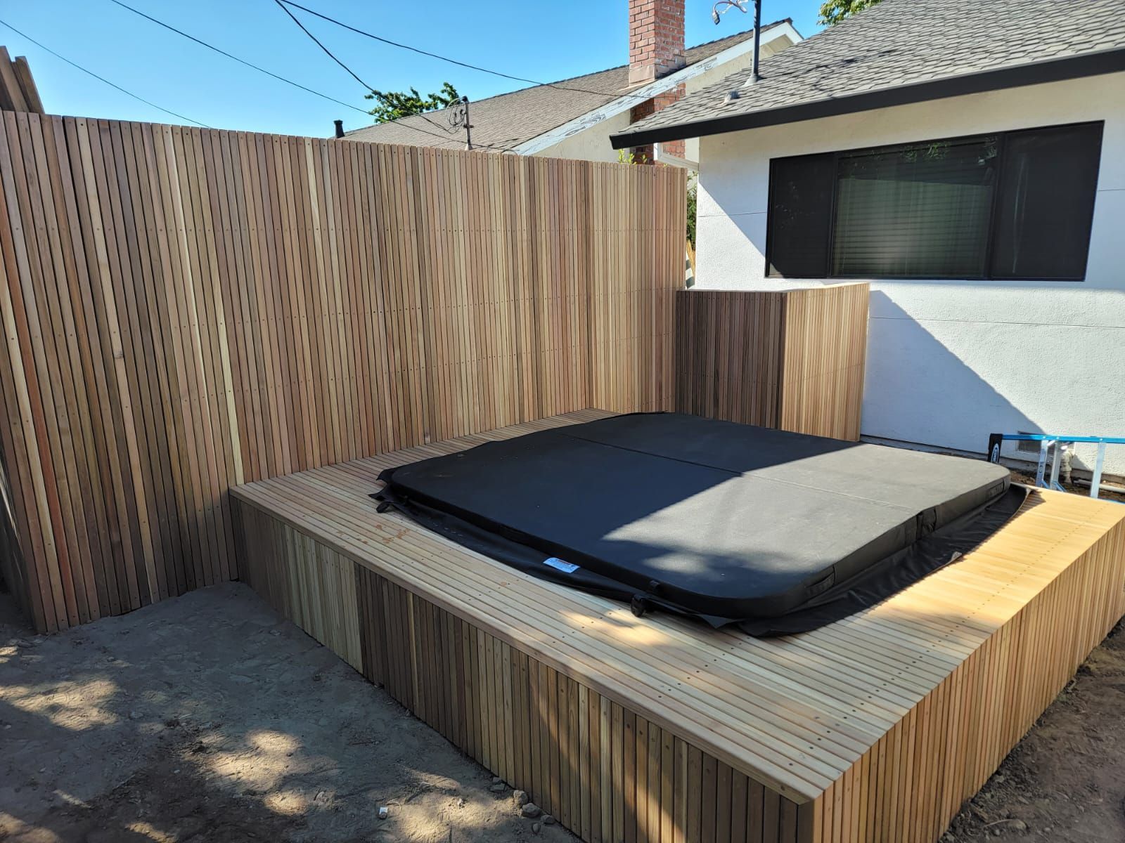 Hot tub with black cover on a wooden deck, enclosed by wooden privacy fence, in a backyard.