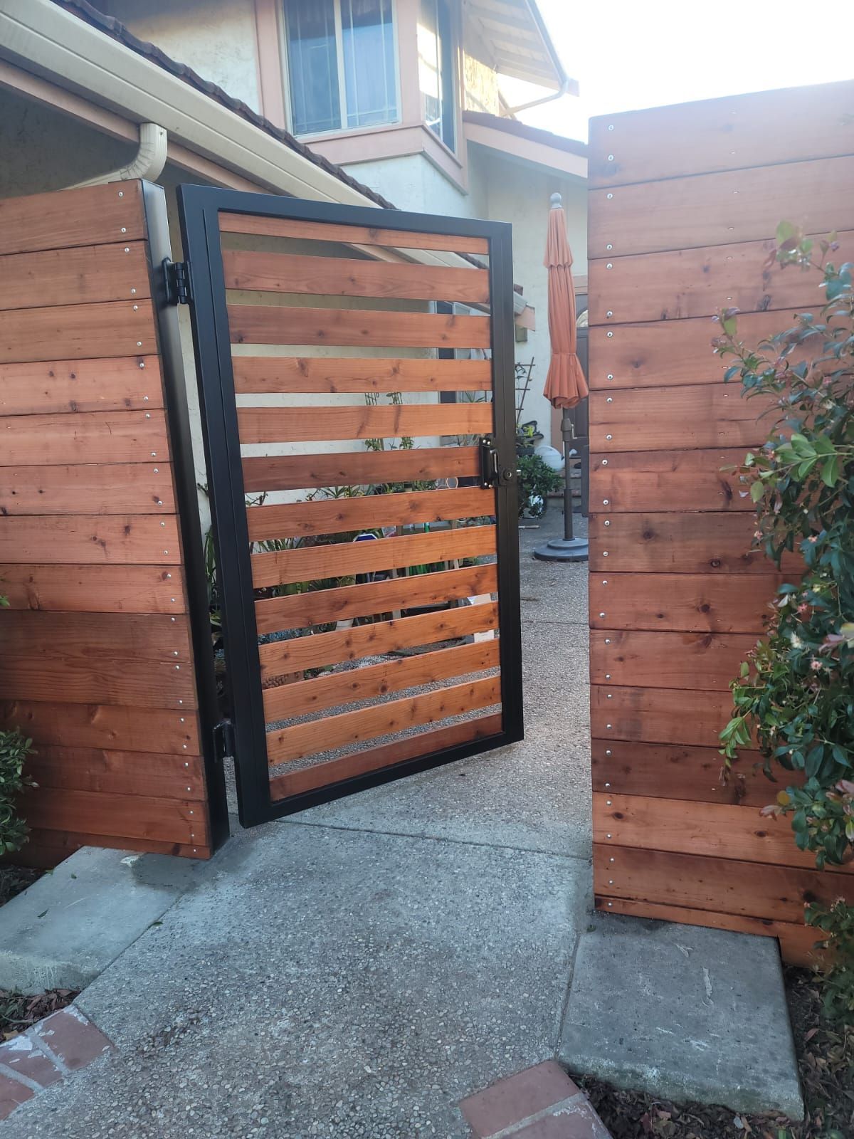 Wooden gate with horizontal slats, flanked by wood fence sections, open on concrete pathway.