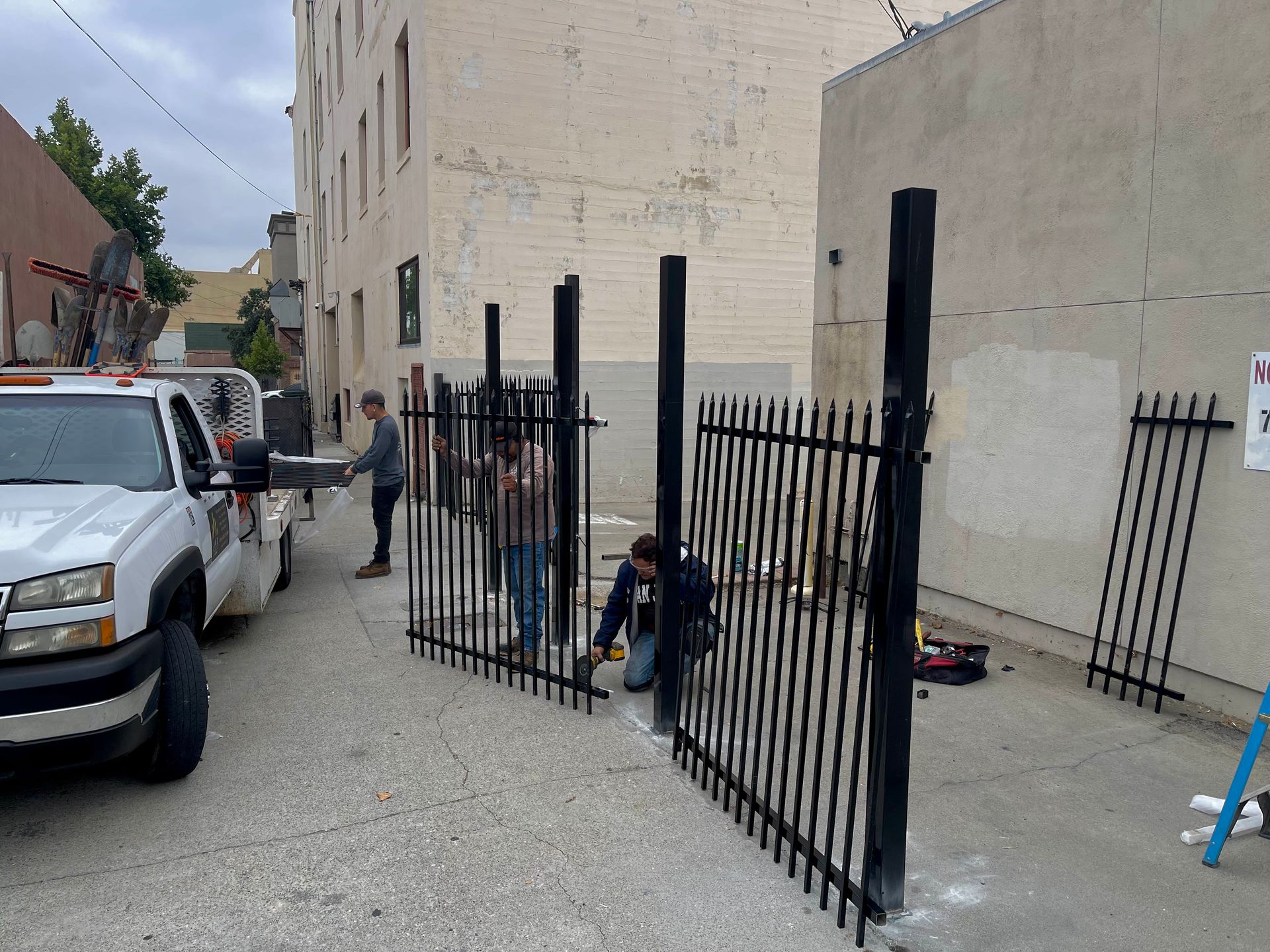 People installing a black metal fence in an alley, near a white truck and building.