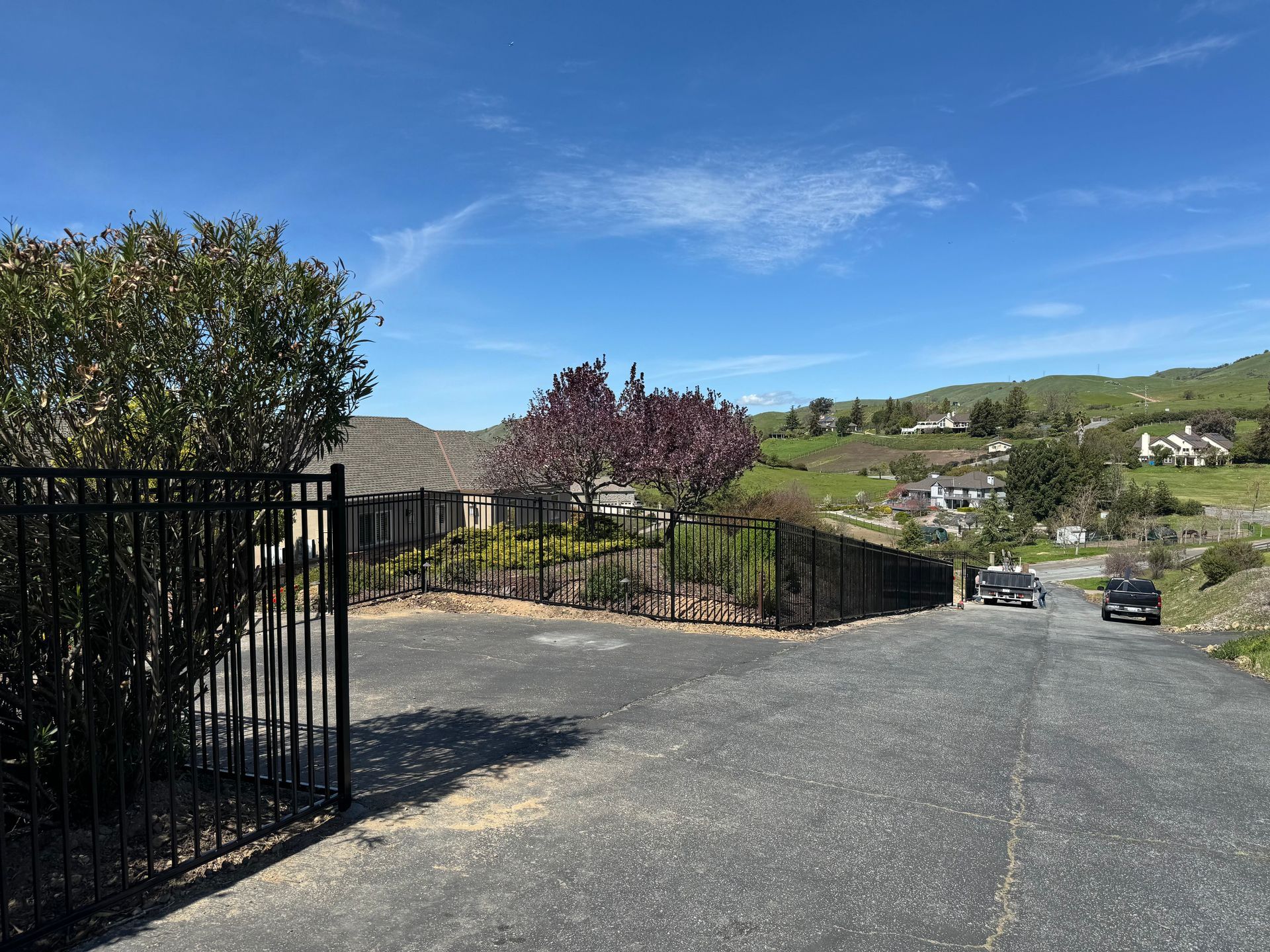 Gated driveway with black fence, trees, houses on hillside, and a bright blue sky.