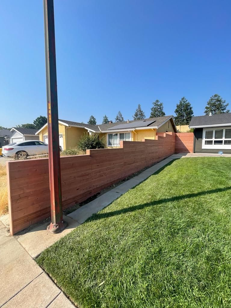 A brown wooden fence separates a green lawn from homes on a sunny day. A utility pole is in the foreground.