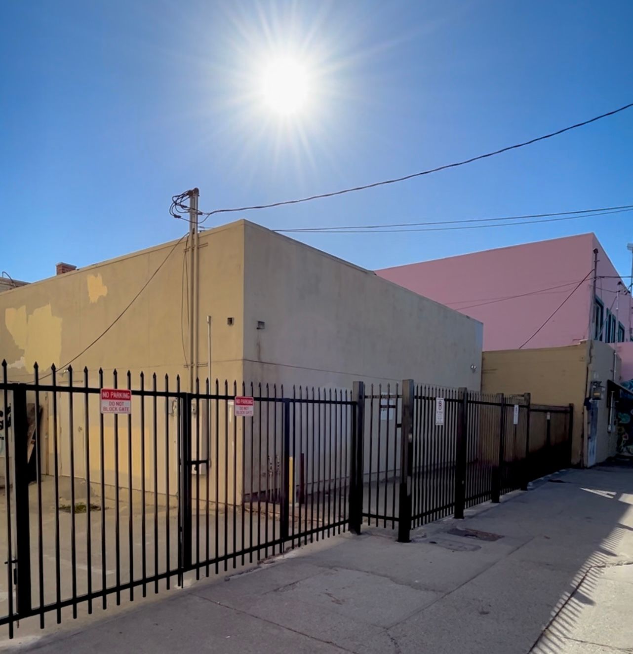 Yellow building behind a black metal fence on a sunny day. Pink building in the background.