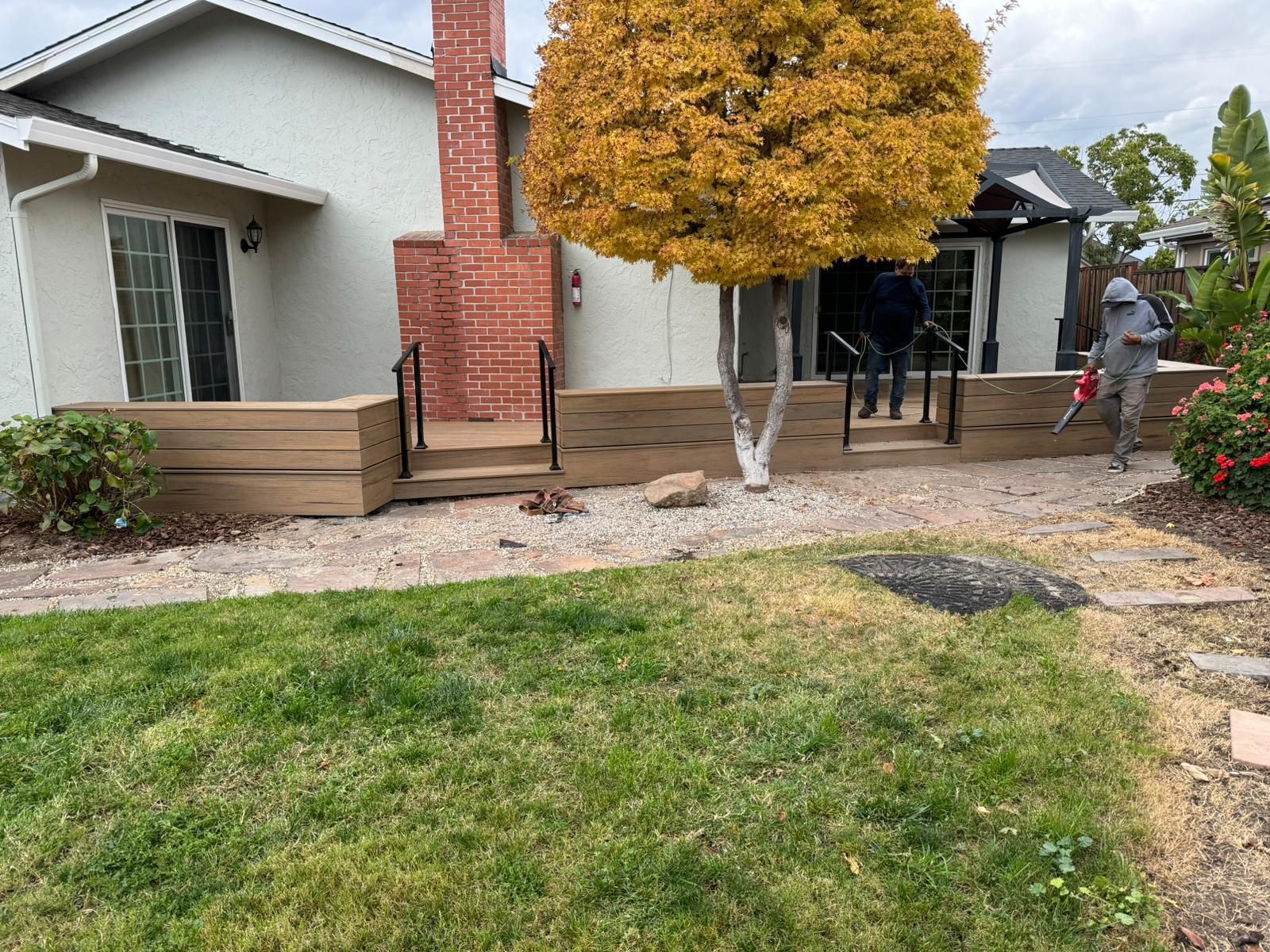 Backyard with a deck and landscaping. A person uses a leaf blower. House and tree in background.