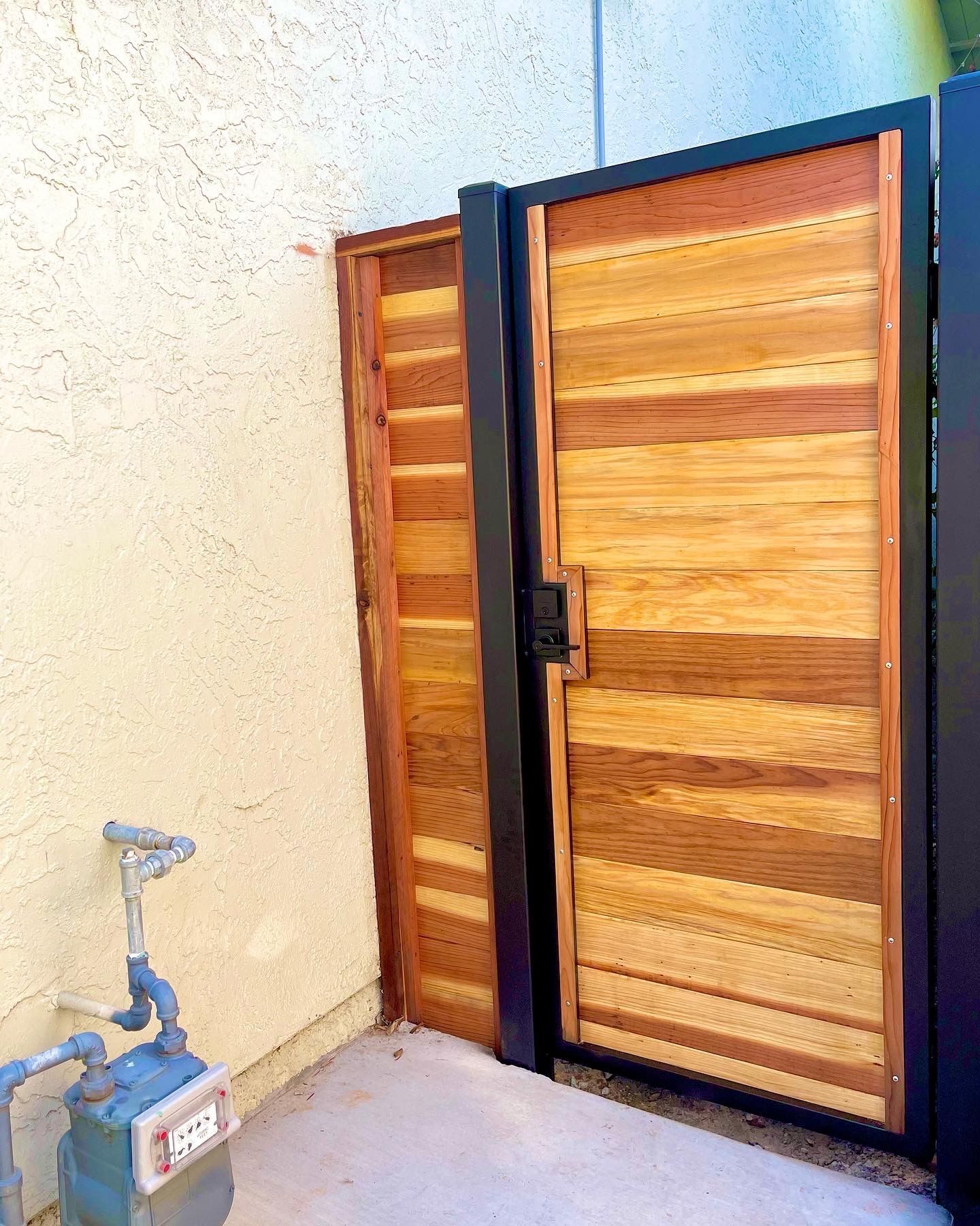 Wooden gate with horizontal planks, black frame, next to stucco wall, outdoor setting.