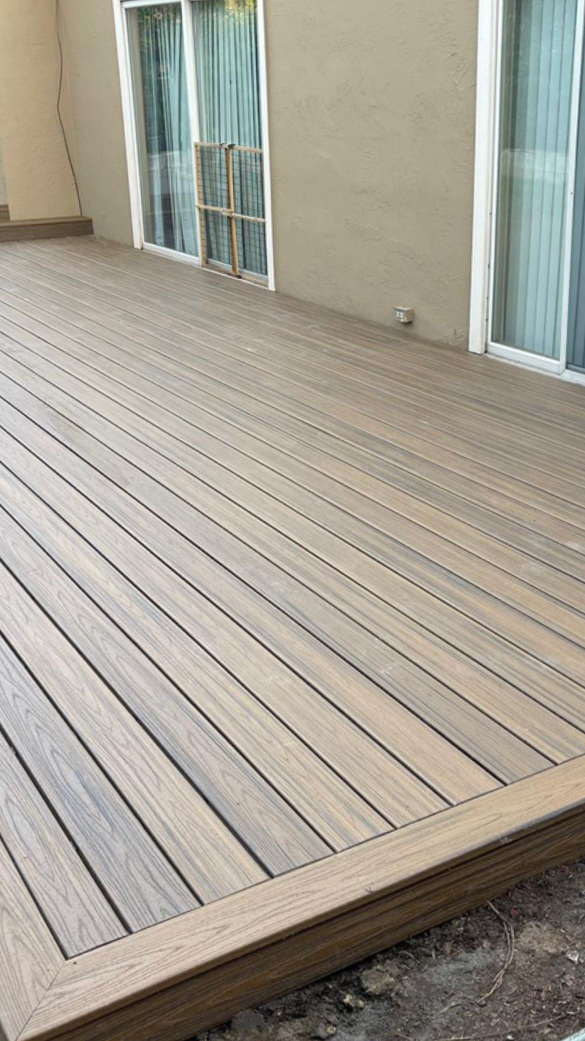 Wooden deck with light-colored planks, bordered by a brown frame, next to a building with windows.