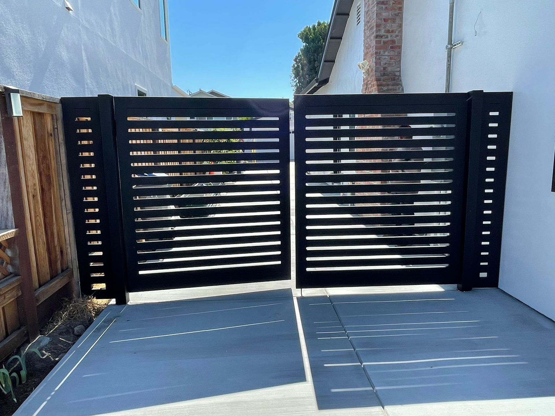 Black slat gates open on a concrete driveway, flanked by white and wooden walls, under a clear sky.