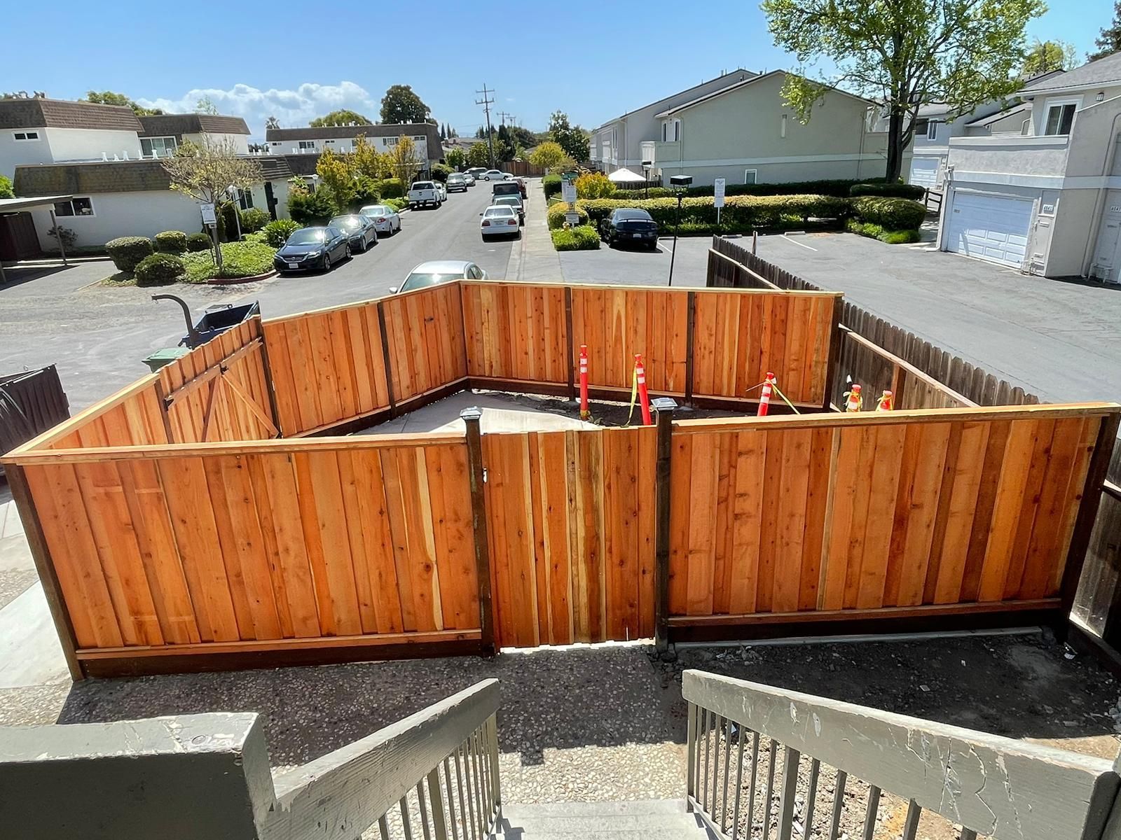 Wooden fence encloses a paved area with utility pipes. Street and houses visible in the background.
