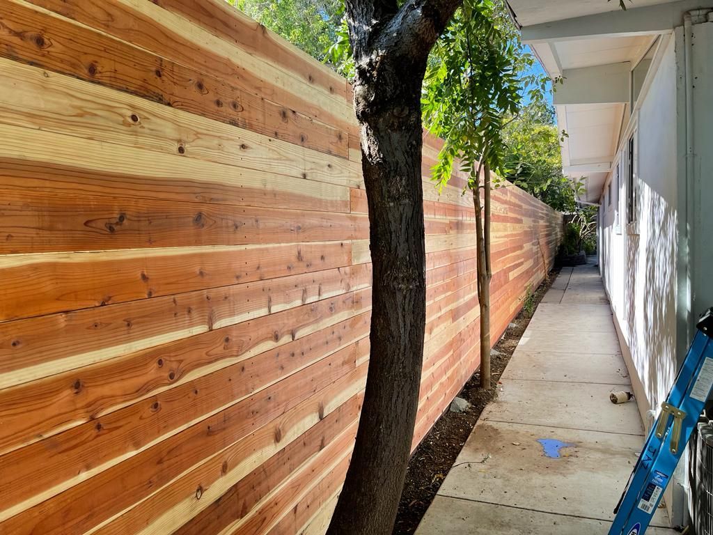 Wooden fence alongside a sidewalk, with a tree in the foreground.