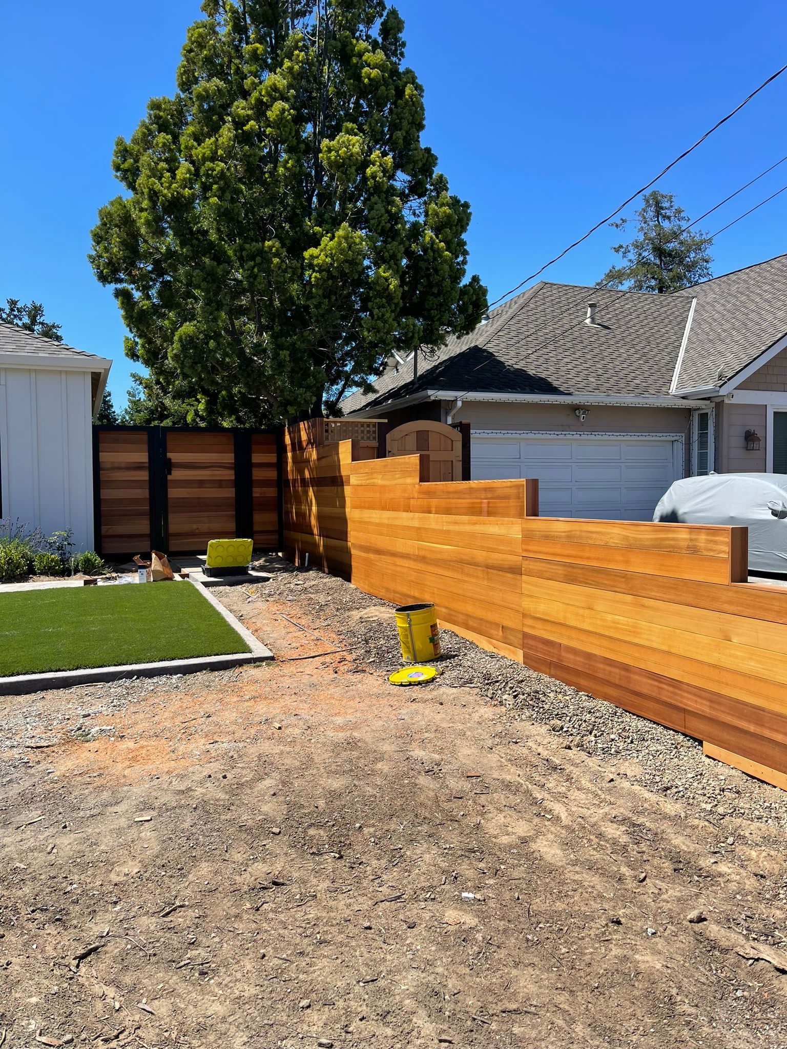 A horizontal cedar fence runs alongside a gravel path and a patch of artificial turf next to a house under a clear sky.