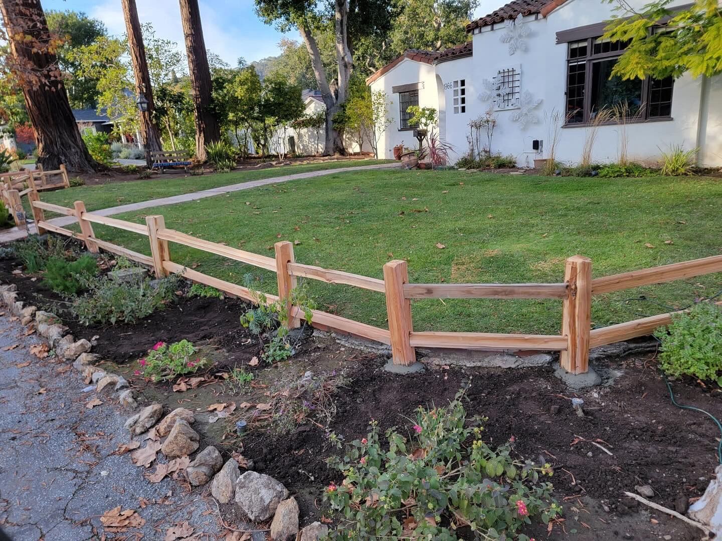 Wooden split-rail fence borders a garden bed in front of a white stucco house with a green lawn.