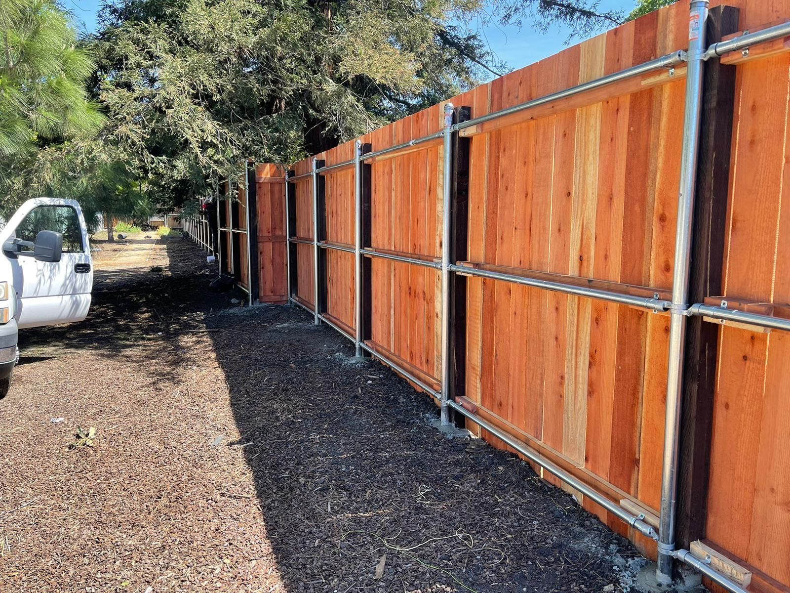 A new wooden fence with metal posts stands along a path, with a white truck parked nearby under trees.