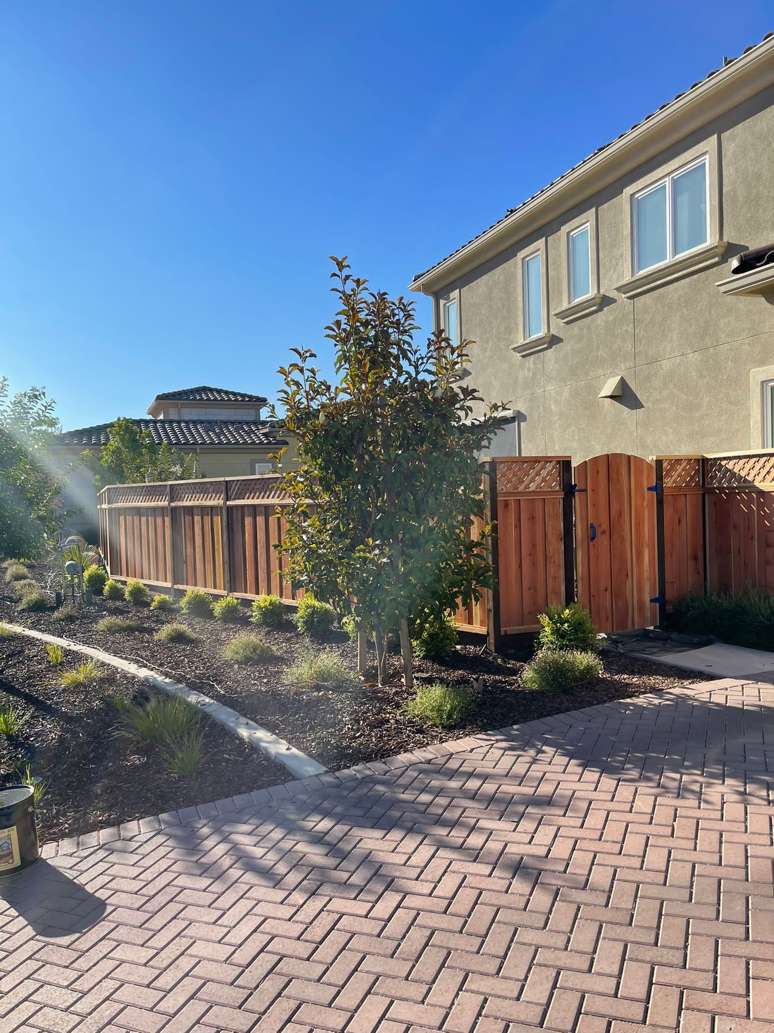 Backyard scene with a wooden fence, brick patio, and a two-story beige house under a clear blue sky.