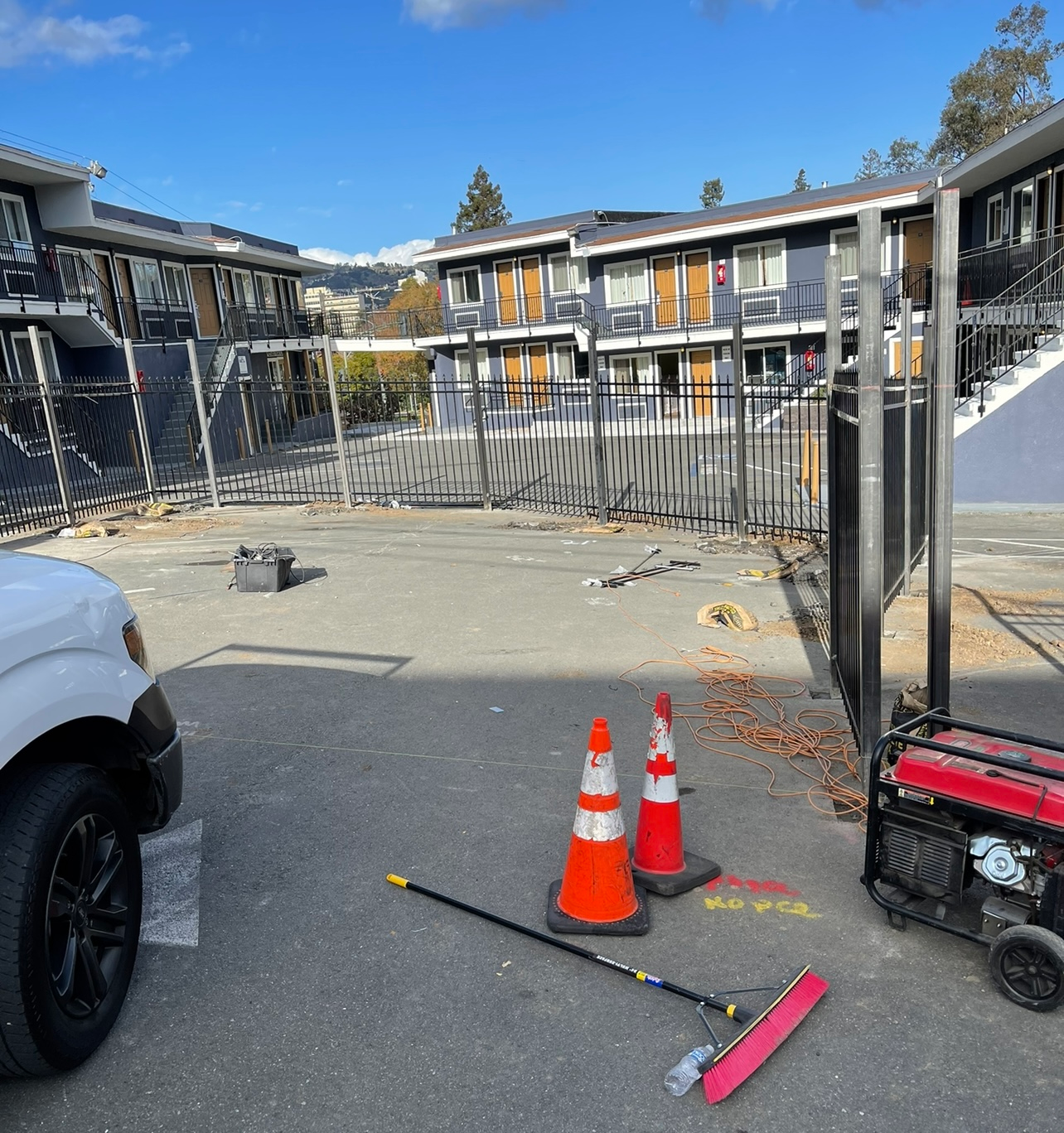 Construction site: car, cones, generator, buildings in background, fence. Blue sky.