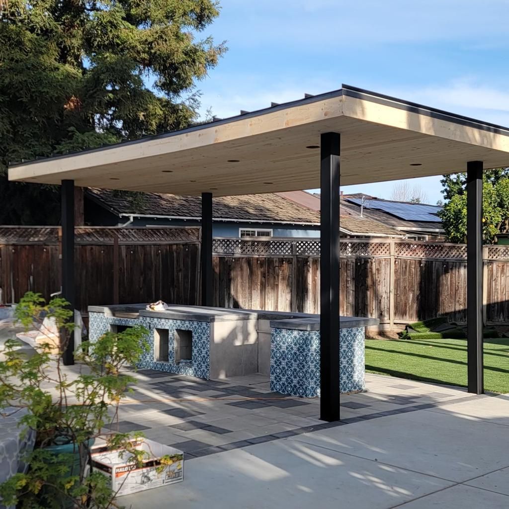 Outdoor kitchen with tile counters, covered by a wood-trimmed pergola, set on a stone patio.