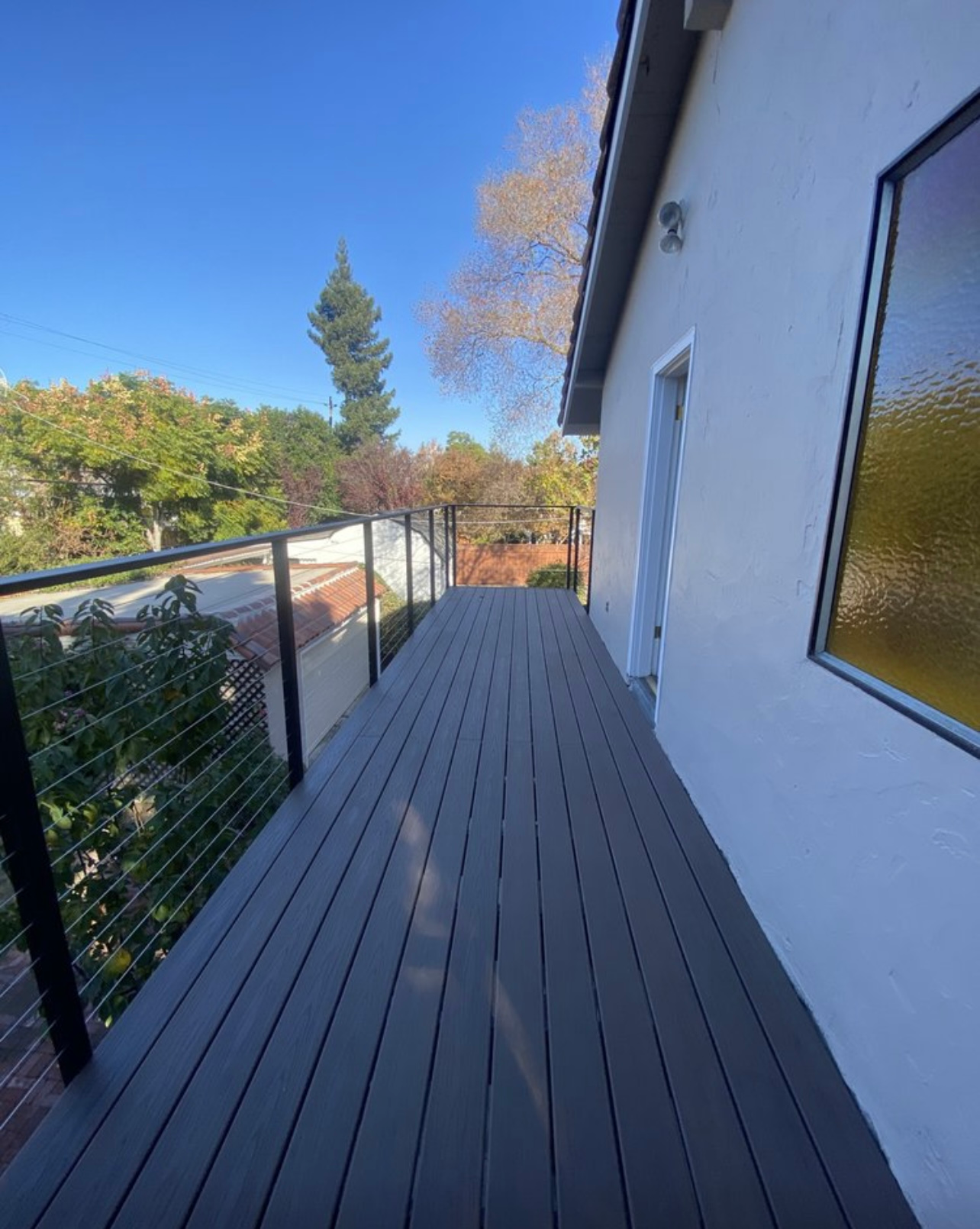 A gray deck with cable railings extends from a white house; a blue sky and trees are in the background.